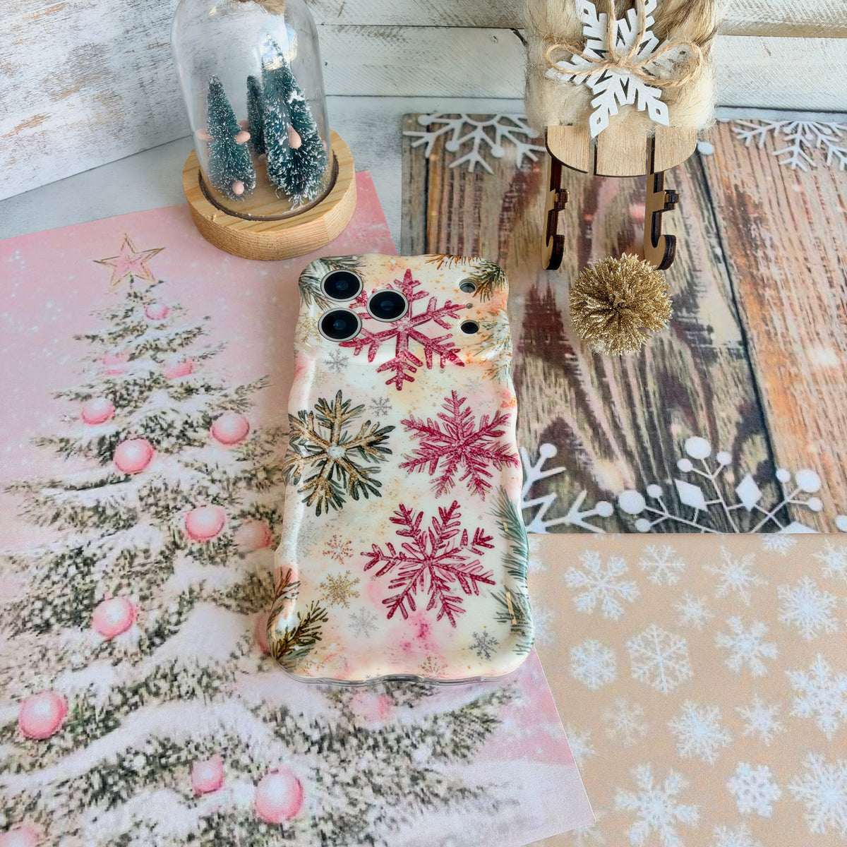 Champagne Snowfall iPhone case lying on whitewashed wood beside a snowflake doily, a cloche of bottle-brush trees and a wooden sleigh