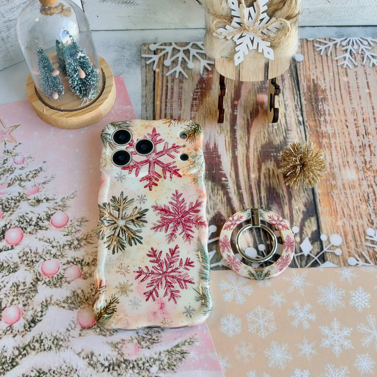 Champagne Snowfall grip ring lying beside the matching pink and gold snowflake iPhone case on whitewashed wood with a wooden sleigh