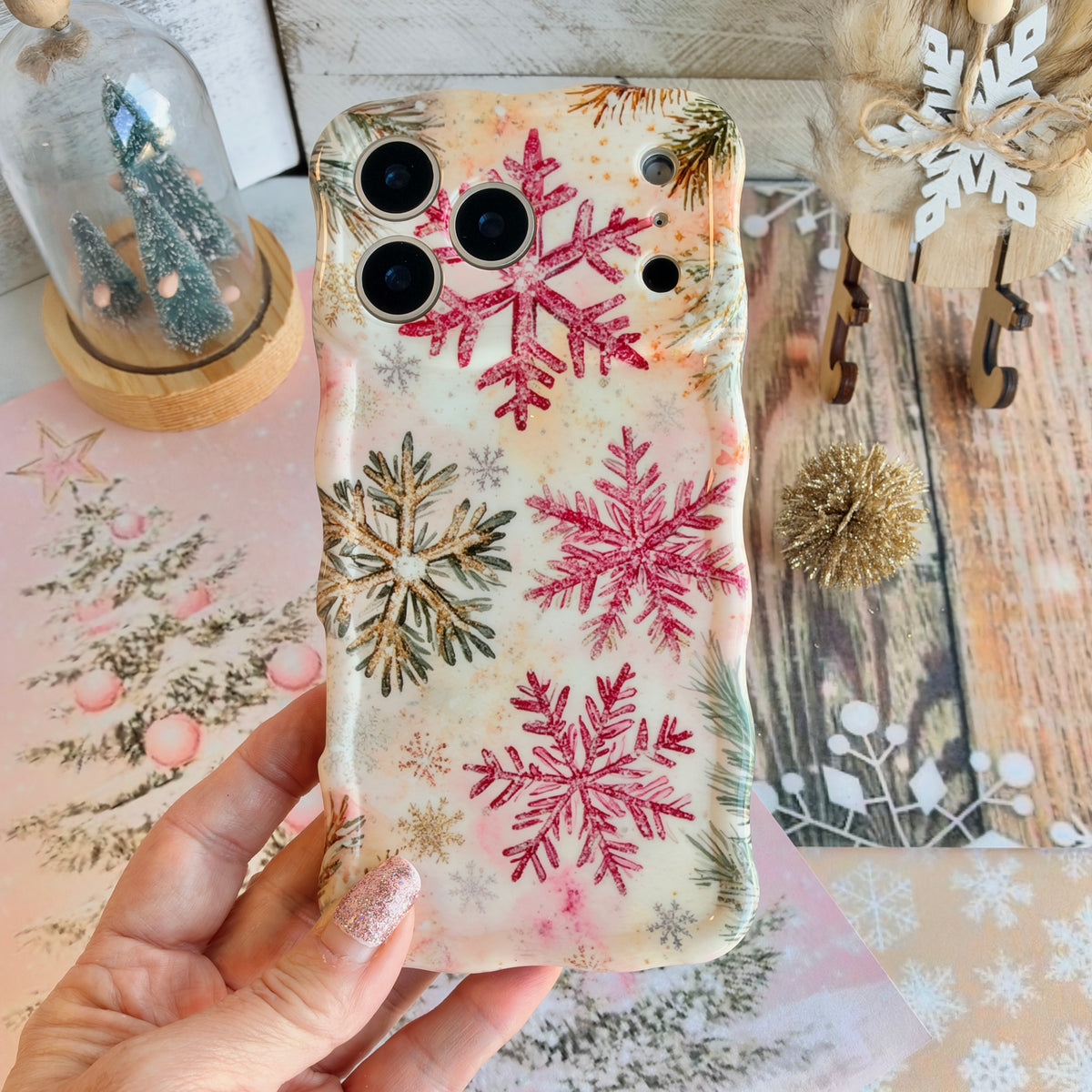 Hand holding the Champagne Snowfall Wavy Luxe MagSafe iPhone case, pink and gold glitter snowflakes, over a pink snowy-pine backdrop
