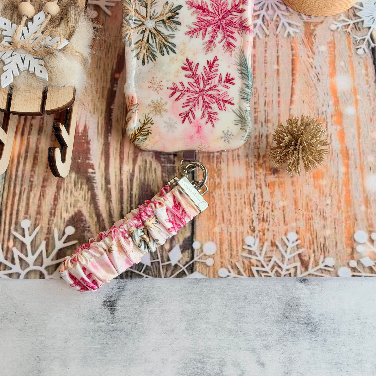 Champagne Snowfall scrunchie phone wrist strap below the matching snowflake iPhone case on whitewashed wood with a snowflake doily