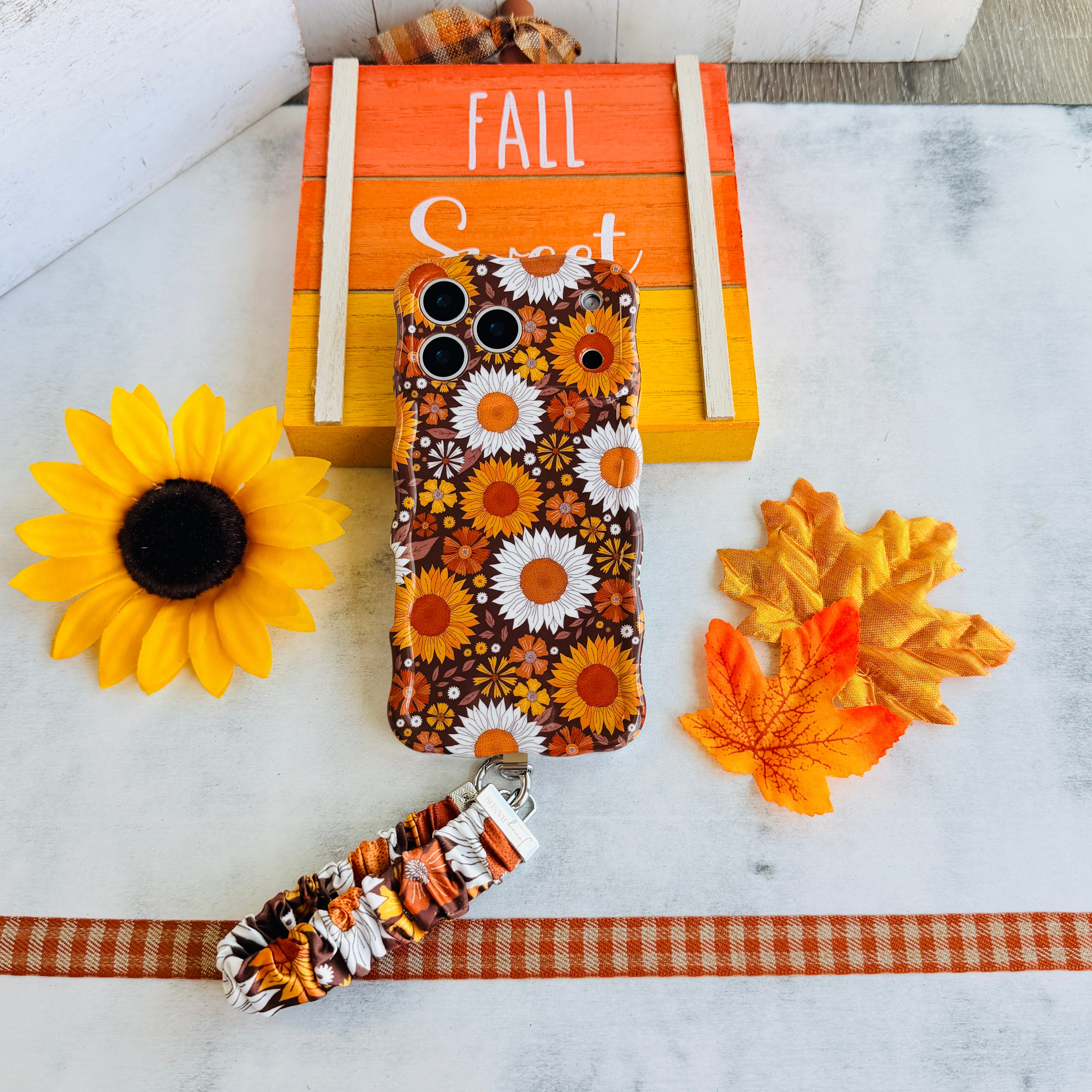 Harvest Sunflowers scrunchie phone wrist strap attached to the matching Harvest Sunflowers iPhone case, photographed against an orange Fall wood sign with a sunflower and maple leaves.
