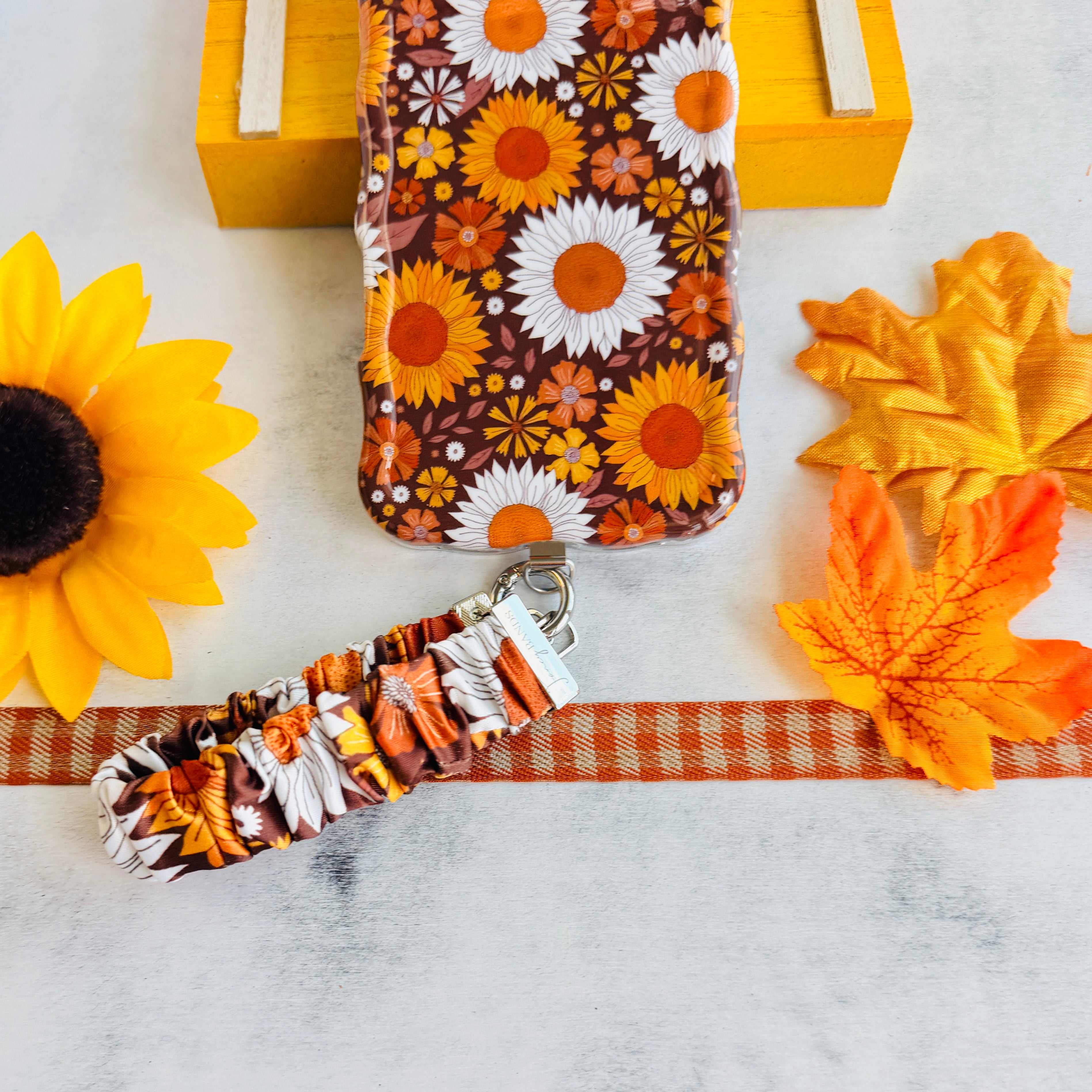 Harvest Sunflowers scrunchie phone wrist strap clipped to the matching Harvest Sunflowers Wavy Luxe iPhone case, styled on orange gingham ribbon with a sunflower, autumn maple leaves, and a yellow wooden crate.