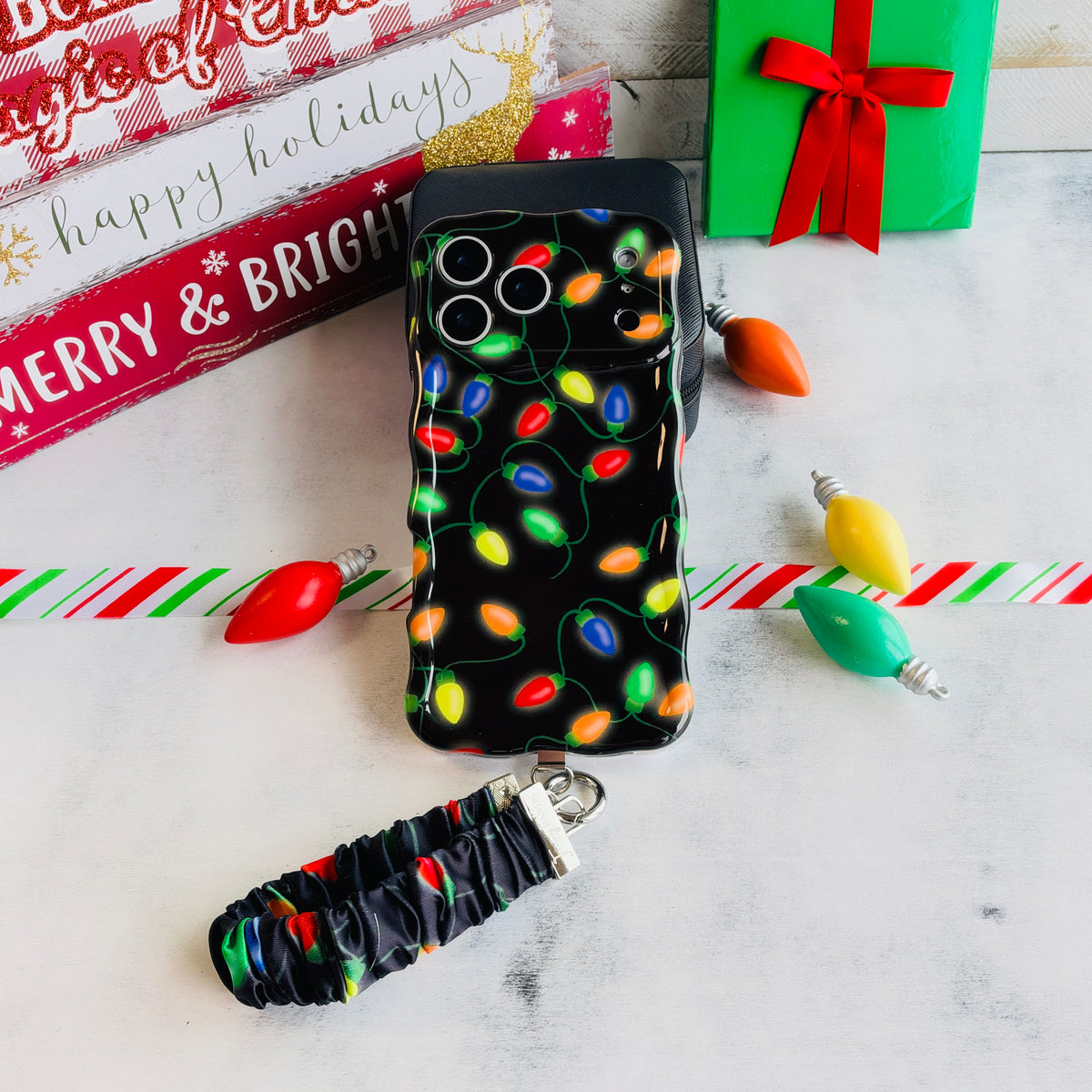 Christmas Lights iPhone case with its black scrunchie wrist strap beside Merry & Bright and happy holidays signs and a green gift