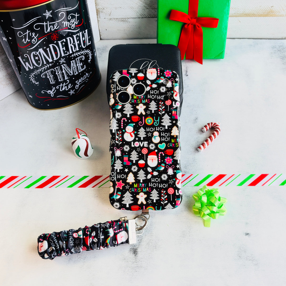 Jolly Wrist-mas iPhone case with its black Christmas icon scrunchie strap beside a Most Wonderful Time of the Year tin and a green gift