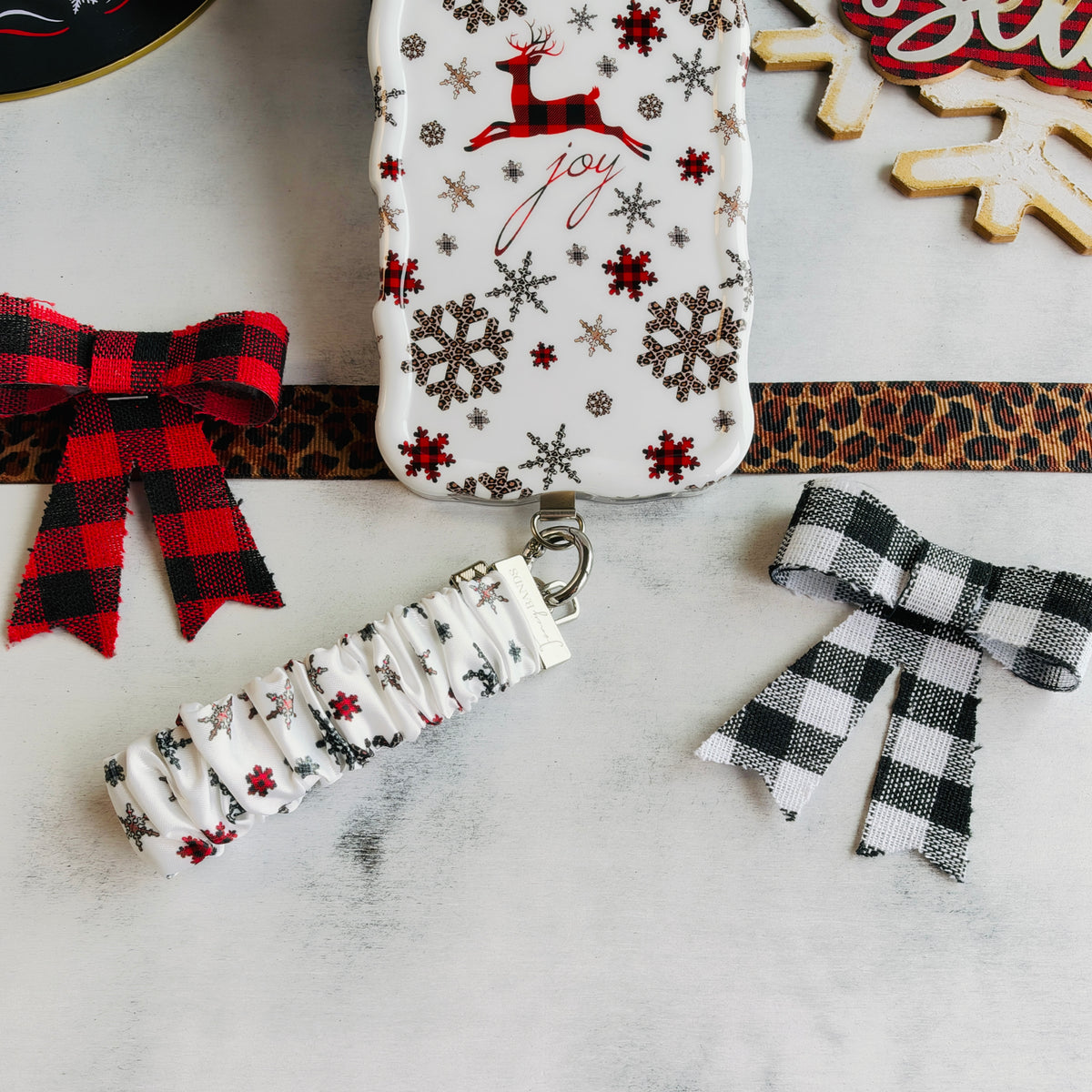 Deer Joy White scrunchie phone wrist strap clipped below the matching reindeer and snowflake iPhone case beside buffalo plaid bows