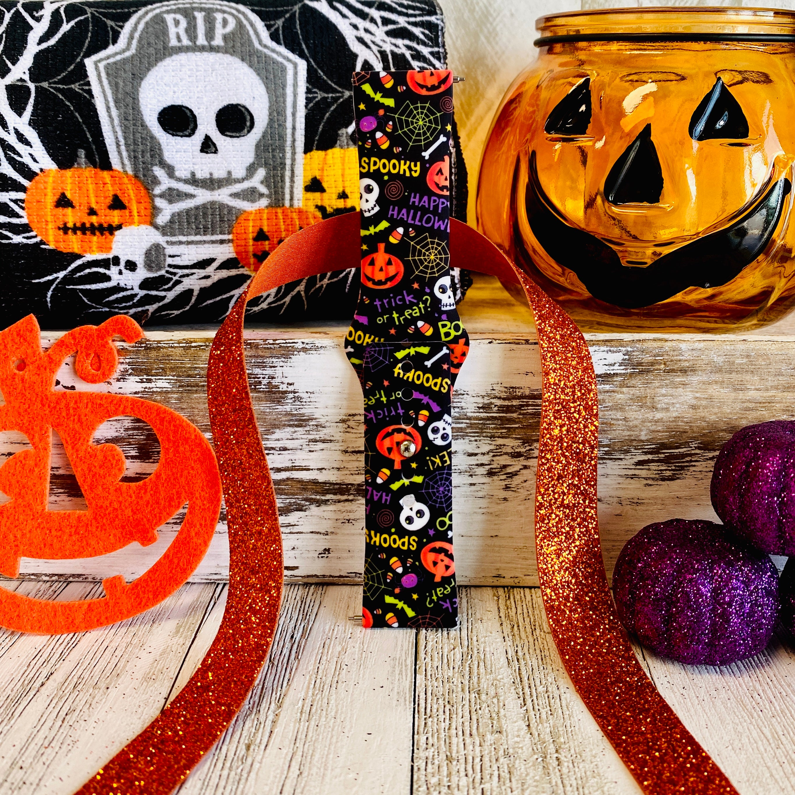 Single Spooky Samsung Galaxy Watch band strap standing in front of a skull tombstone with purple and orange felt pumpkins.