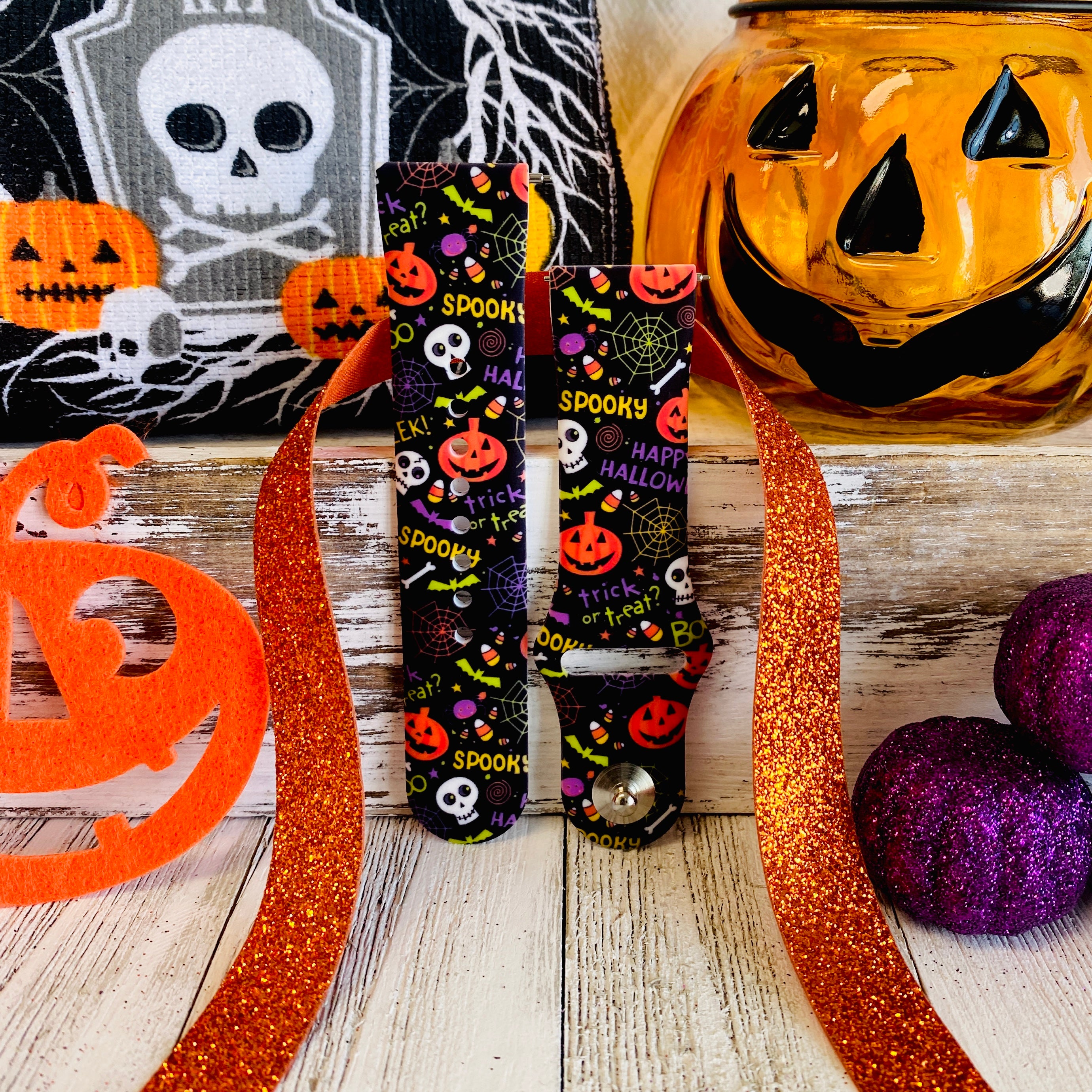 Two Spooky Samsung Galaxy Watch band straps standing side by side with orange glitter ribbon and a glowing jack-o-lantern behind.