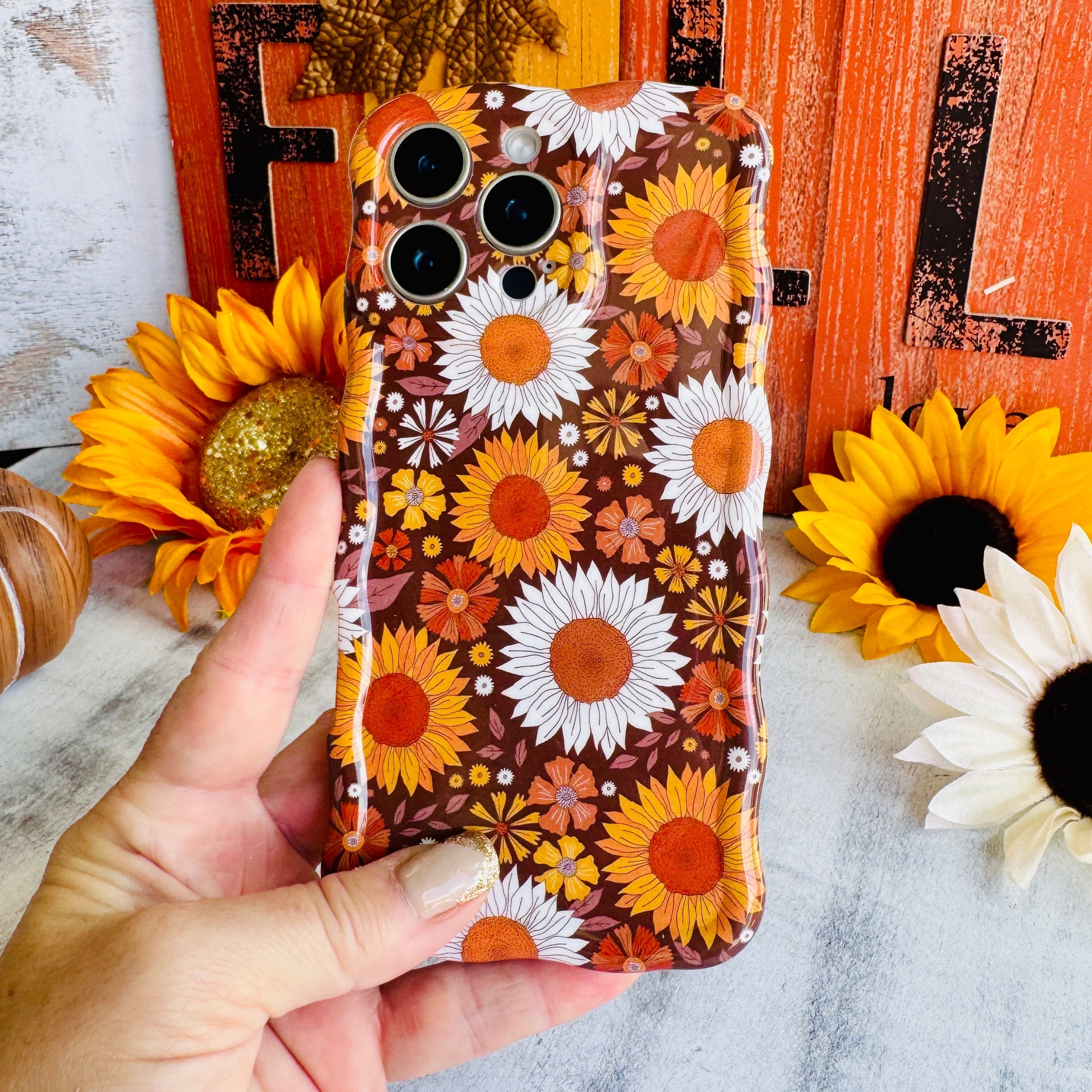 Harvest Sunflowers Wavy Luxe MagSafe iPhone case held against sunflowers and a rust barn door — the Fancy Bands Fall 2026 Collection
