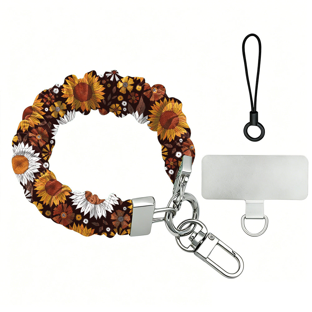 Harvest Sunflowers scrunchie phone wrist strap on a white background, showing the deep brown fabric packed with golden sunflowers and white daisies, silver lobster clasp, black loop cord, and clear attachment tab.