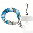 Blue and green satin Beach Please scrunchie phone wrist strap clipped to the matching Beach Please Wavy Luxe iPhone case with a beach chair and cocktail design, styled on a sandy shoreline background with starfish and ocean waves.