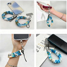Blue and green satin Beach Please scrunchie phone wrist strap clipped to the matching Beach Please Wavy Luxe iPhone case with a beach chair and cocktail design, styled on a sandy shoreline background with starfish and ocean waves.