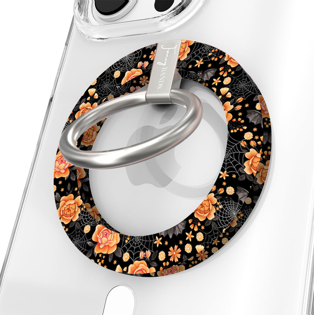 Haunted Blossoms Hold It Fancy MagSafe grip ring close-up on a clear iPhone case, showing the black ring with burnt-orange roses, mushrooms and gold spiderwebs around the silver rotating ring.