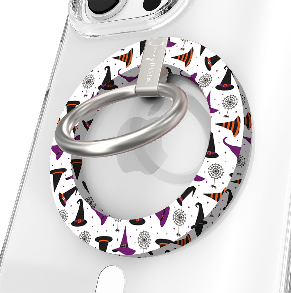 Witch Please Hold It Fancy MagSafe grip ring close-up on a clear iPhone case, showing the white ring scattered with purple, black and striped orange witch hats around the silver rotating ring.