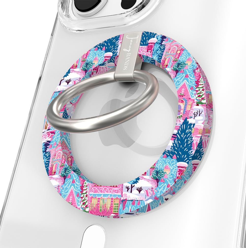 Close-up of the Sugarplum Square MagSafe grip ring with pink gingerbread houses and blue pines and a brushed silver ring on a clear iPhone case