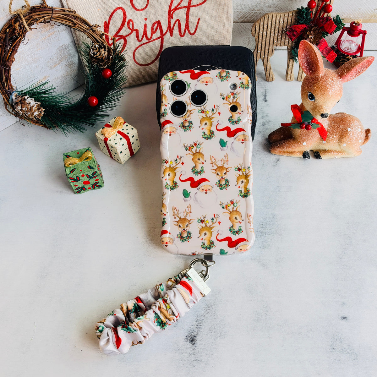 Vintage Christmas iPhone case with its cream scrunchie wrist strap beside a holly wreath, a vintage fawn figurine and a Bright sign