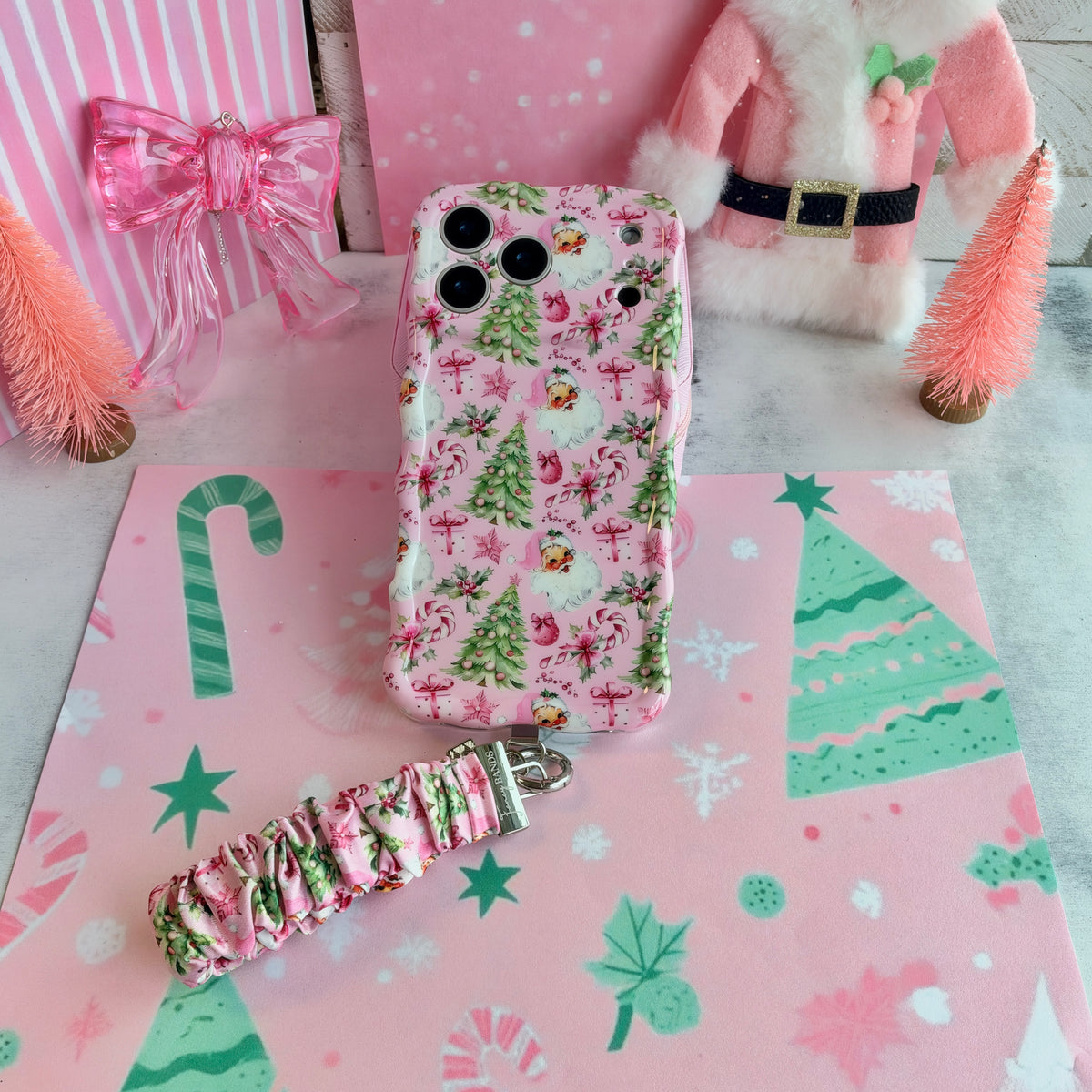 Jolly Pink Santa iPhone case with its scrunchie wrist strap beside a pink satin bow, a pink Santa coat and pink bottle-brush trees