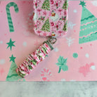 Jolly Pink Santa scrunchie phone wrist strap clipped below the matching iPhone case on pink paper with green Christmas trees