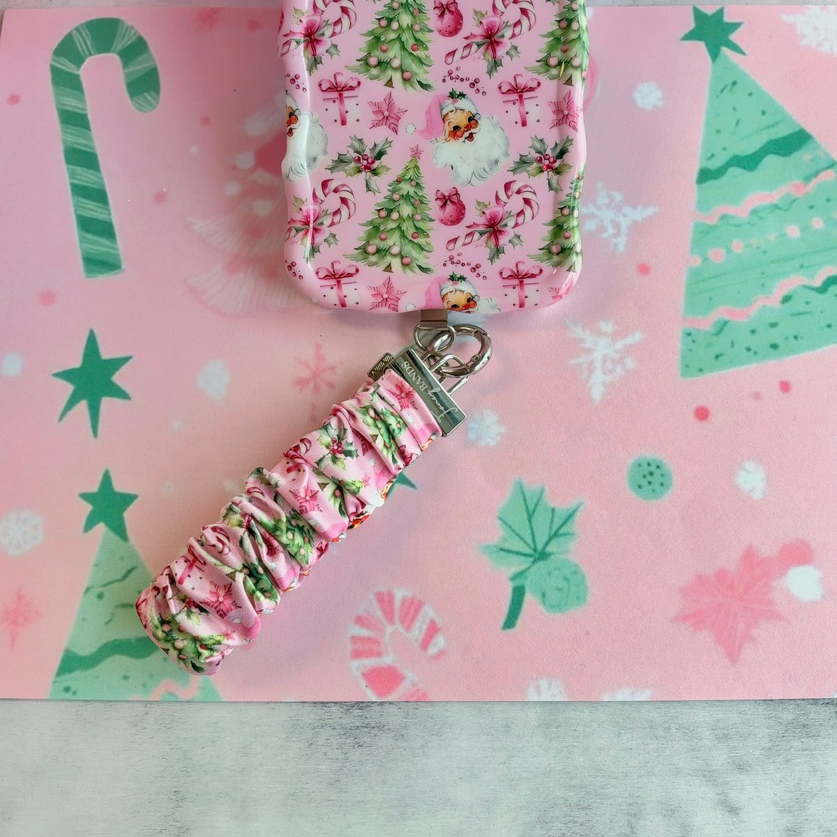Jolly Pink Santa scrunchie phone wrist strap clipped below the matching iPhone case on pink paper with green Christmas trees