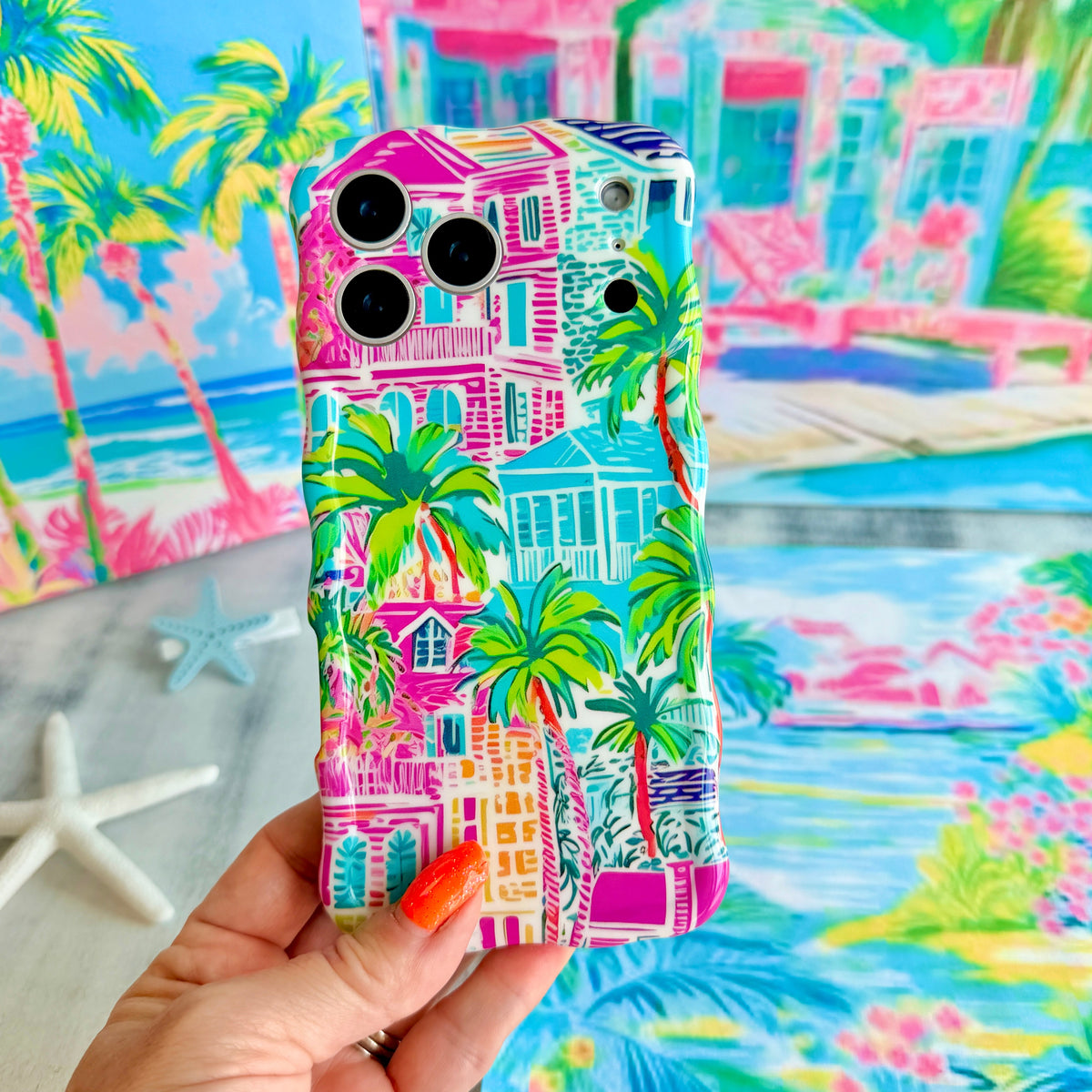 Hand holding the La Palma Villa Wavy Luxe MagSafe iPhone case, painted pink villas and palm trees on a tropical backdrop