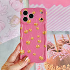 Hand holding the Caramel Coated Cutie Wavy Luxe MagSafe iPhone case, showing the rose-pink base scattered with golden caramel apples against a pink fall mood board backdrop.