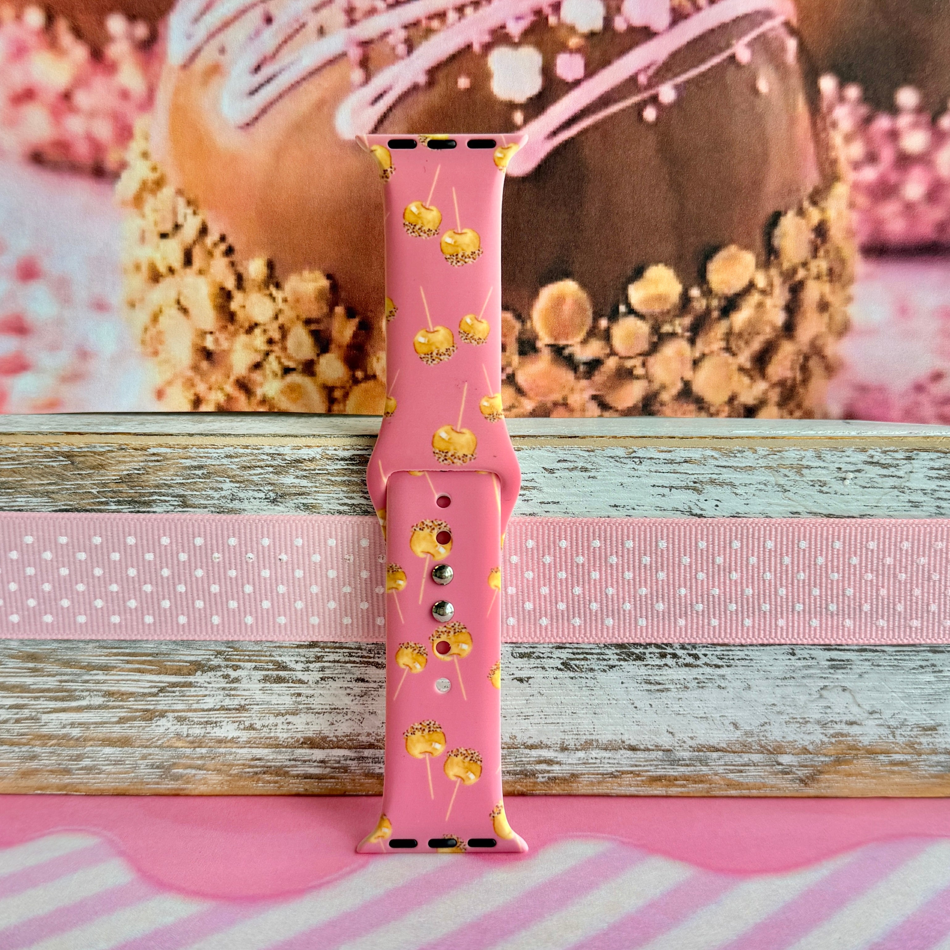 Caramel Coated Cutie Apple Watch band single strap half standing on weathered wood, showing the golden caramel apples on rose-pink silicone.
