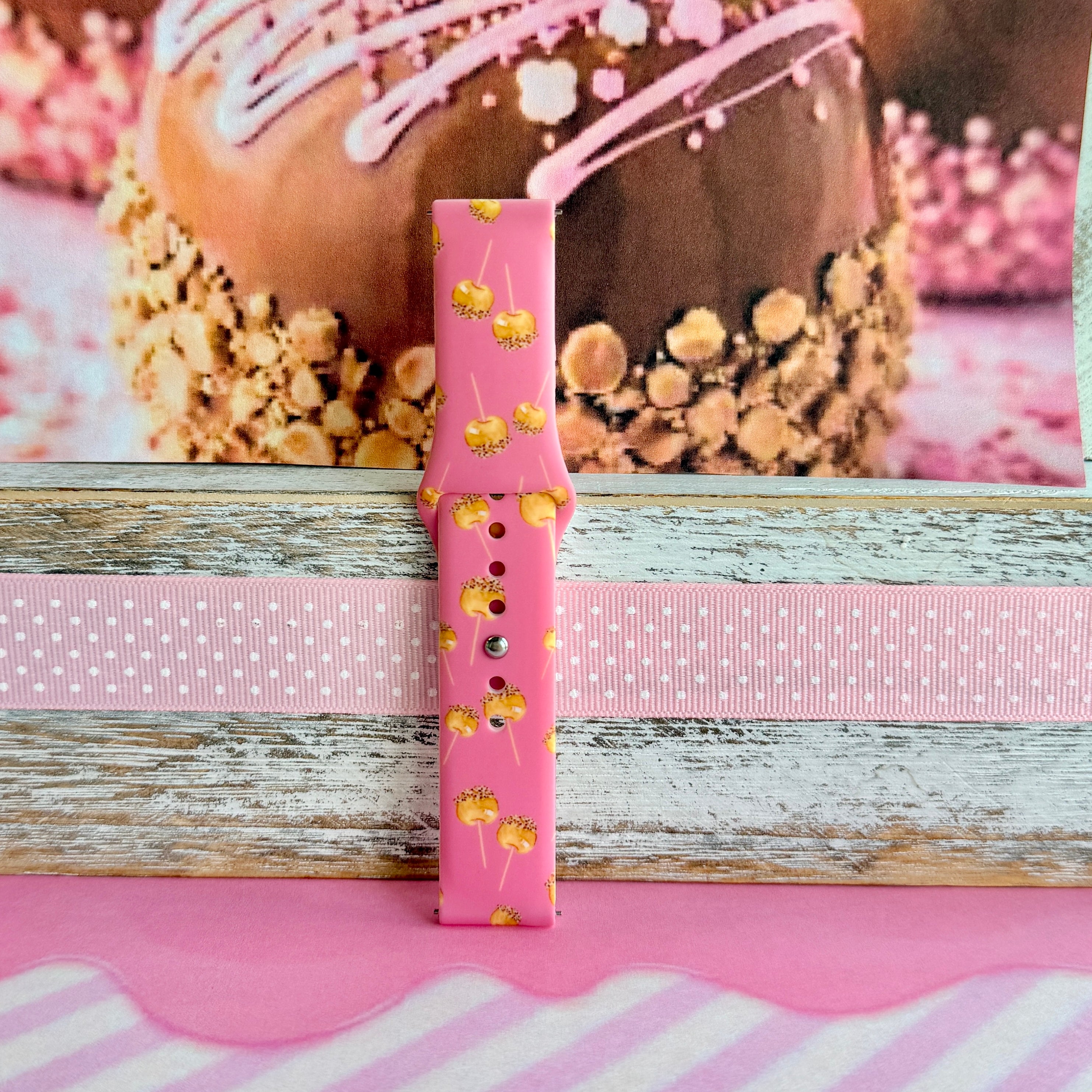 Caramel Coated Cutie Samsung Galaxy Watch band single strap half standing on weathered wood, showing the golden caramel apples on rose-pink silicone.