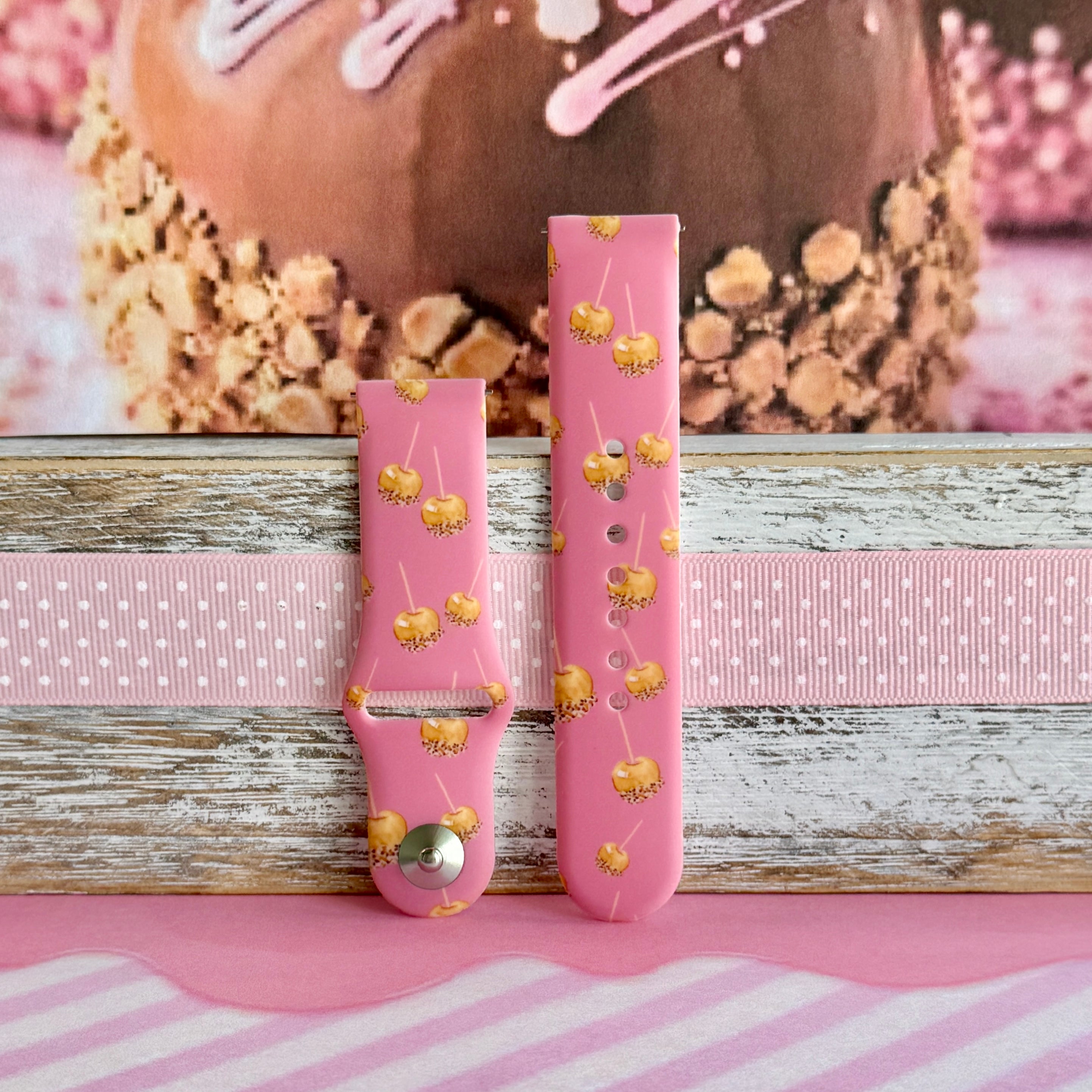 Caramel Coated Cutie Samsung Galaxy Watch band with both strap halves side by side on weathered wood and pink ribbon, showing the full caramel apple print.