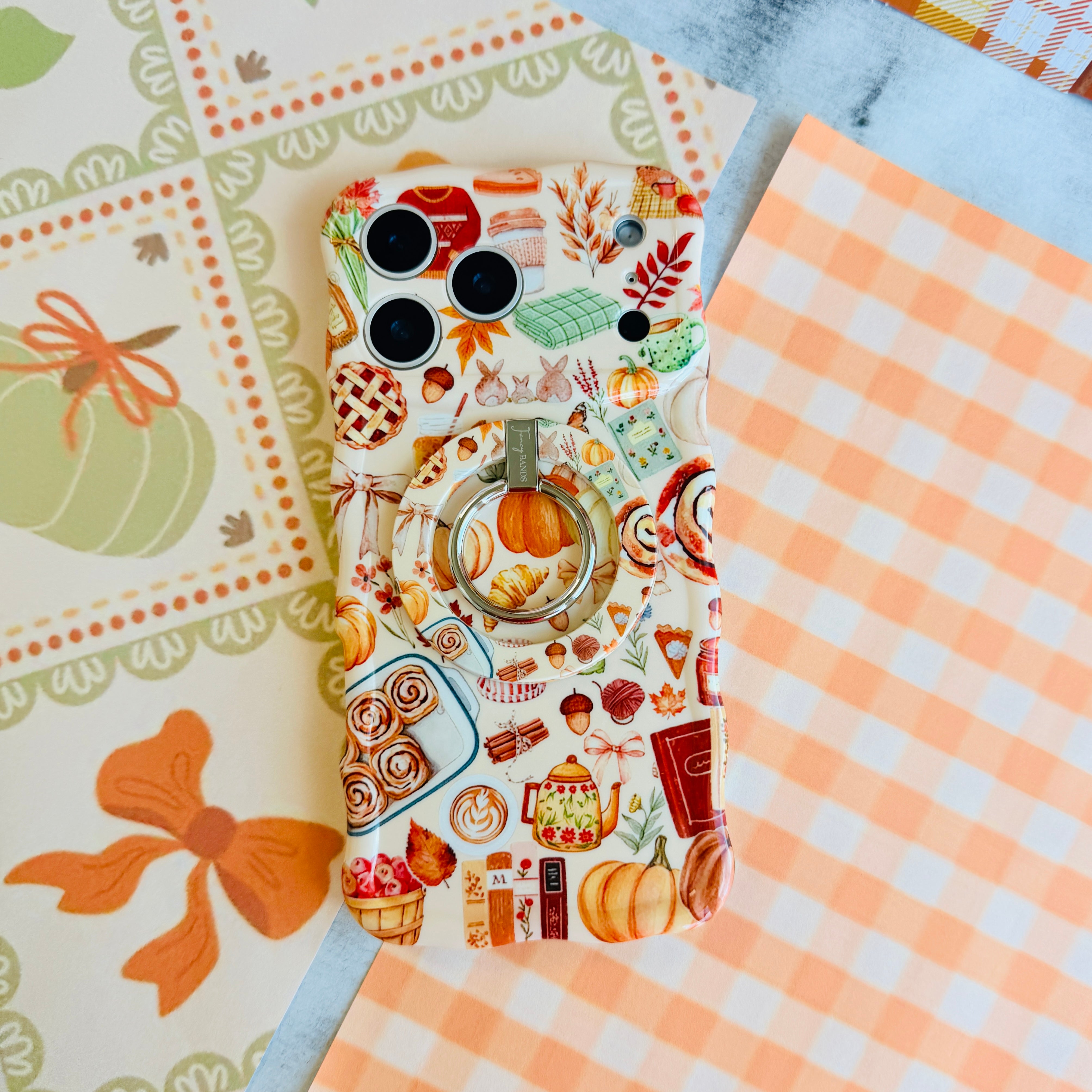 All The Fall Things MagSafe grip ring attached to the matching All The Fall Things iPhone case, styled on orange gingham and a pumpkin illustration backdrop.