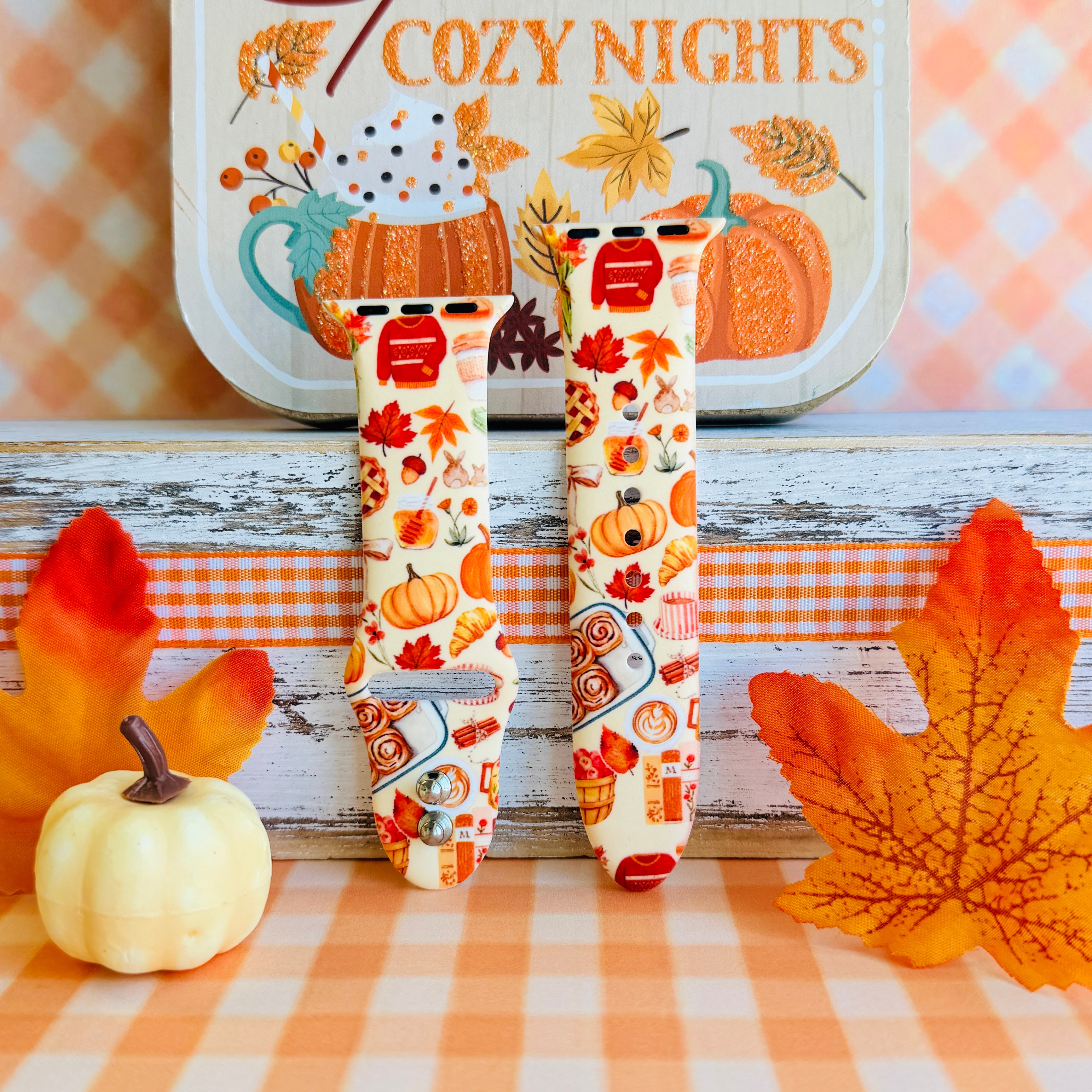 All The Fall Things Apple Watch band with both strap halves standing on weathered wood beside a white pumpkin and orange gingham ribbon, showing the full watercolor collage print.
