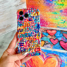 Hand holding the Love Riot Wavy Luxe MagSafe iPhone case with graffiti lettering and painted hearts on a brick wall background