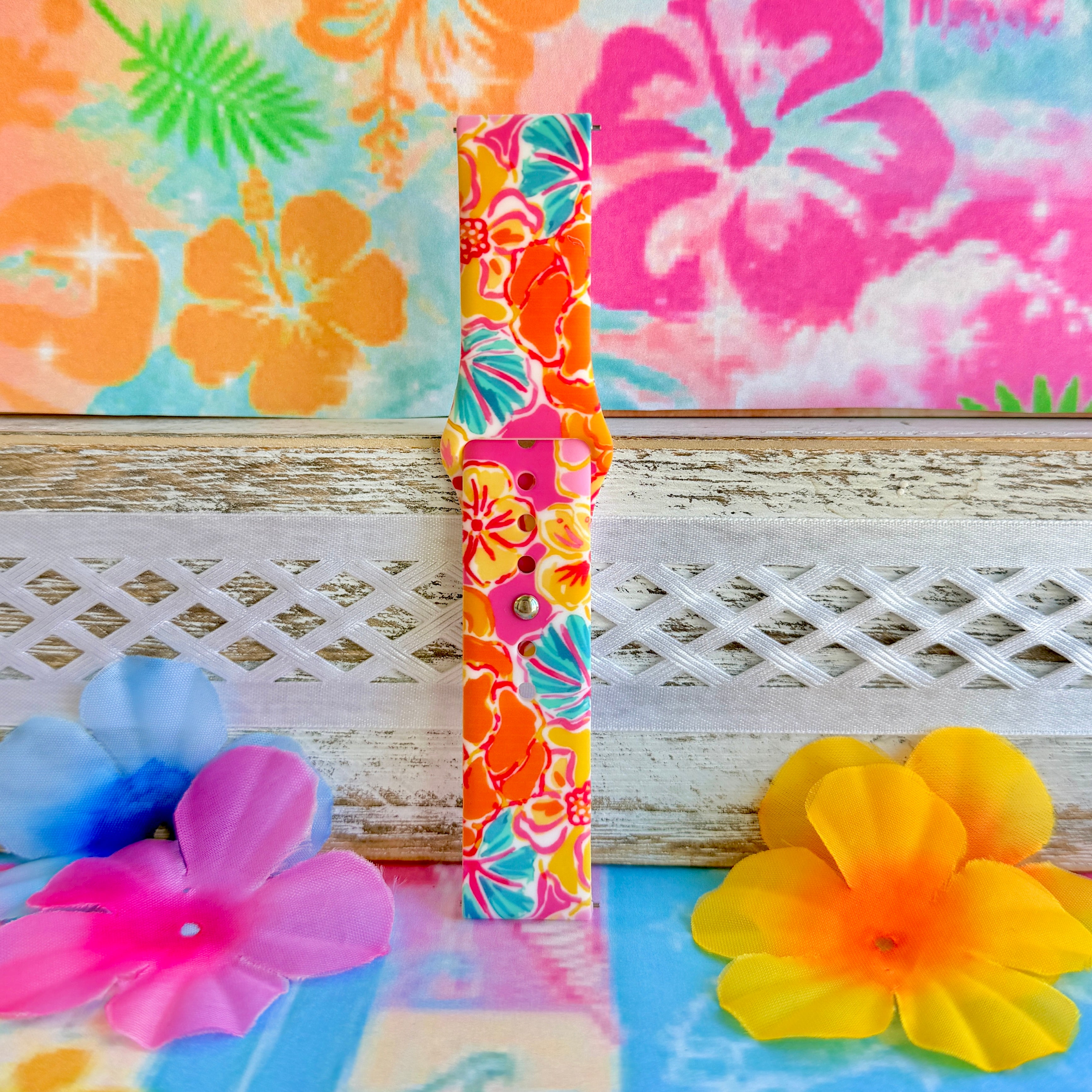 Sunset Hibiscus silicone Samsung Watch band with vibrant pink, orange, yellow, and teal tropical hibiscus floral print, shown displayed and worn on Apple Watch against a colorful tropical floral backdrop.