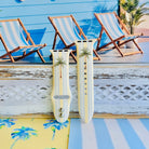 Palm Shore Club silicone Apple Watch band with hand-painted palm trees over soft blue and yellow cabana stripes on an ivory base, shown displayed and worn on Apple Watch against a coastal beach club backdrop.