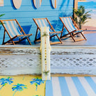 Palm Shore Club silicone Samsung Watch band with hand-painted palm trees over soft blue and yellow cabana stripes on an ivory base, shown displayed and worn on Apple Watch against a coastal beach club backdrop.