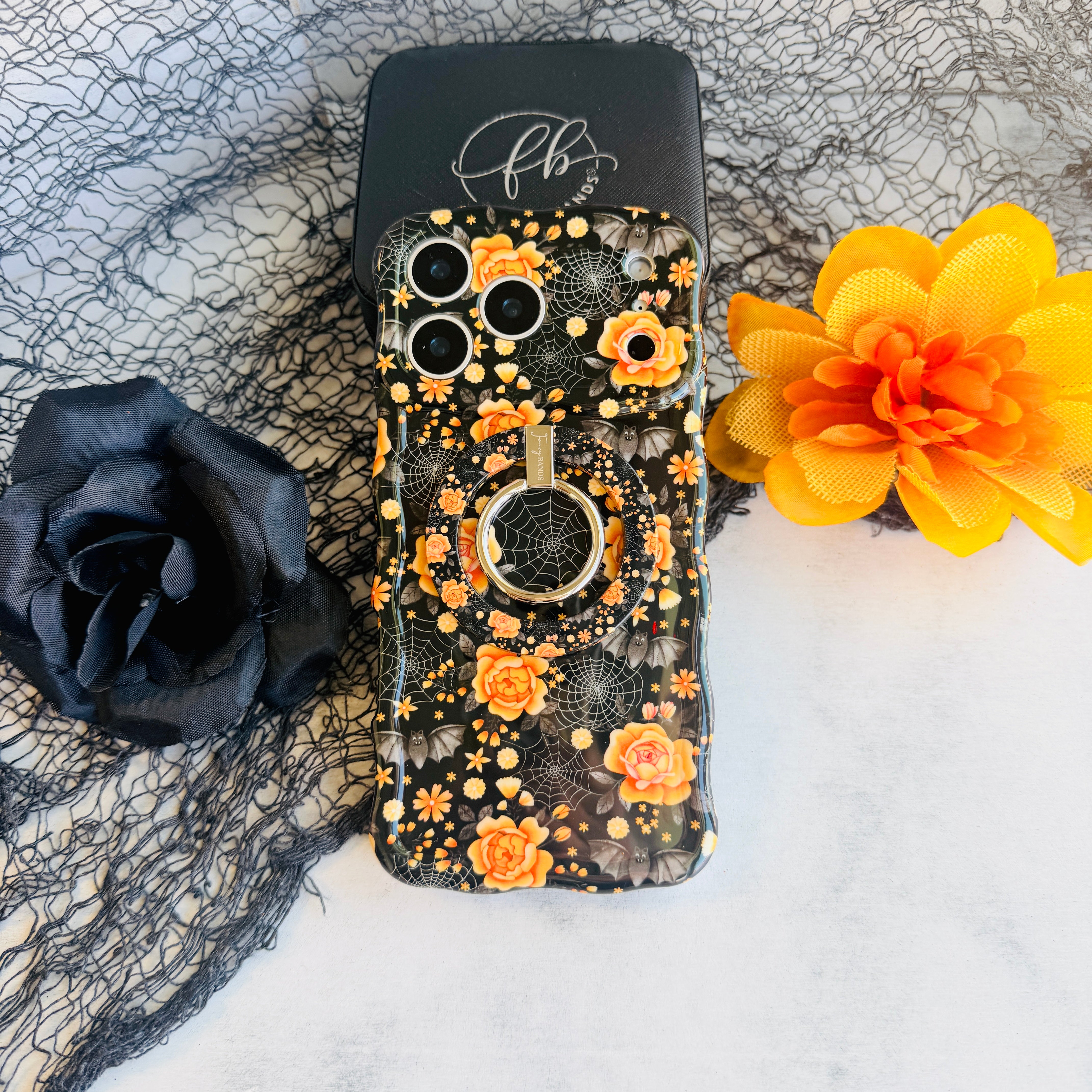 Haunted Blossoms MagSafe grip ring attached to the matching black coral rose Wavy Luxe case, styled with a black rose and an orange bloom.