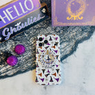 Witch Please MagSafe grip ring attached to the matching cream witch hat Wavy Luxe case, shown with a HELLO Ghoulies sign and Book of Spells backdrop.