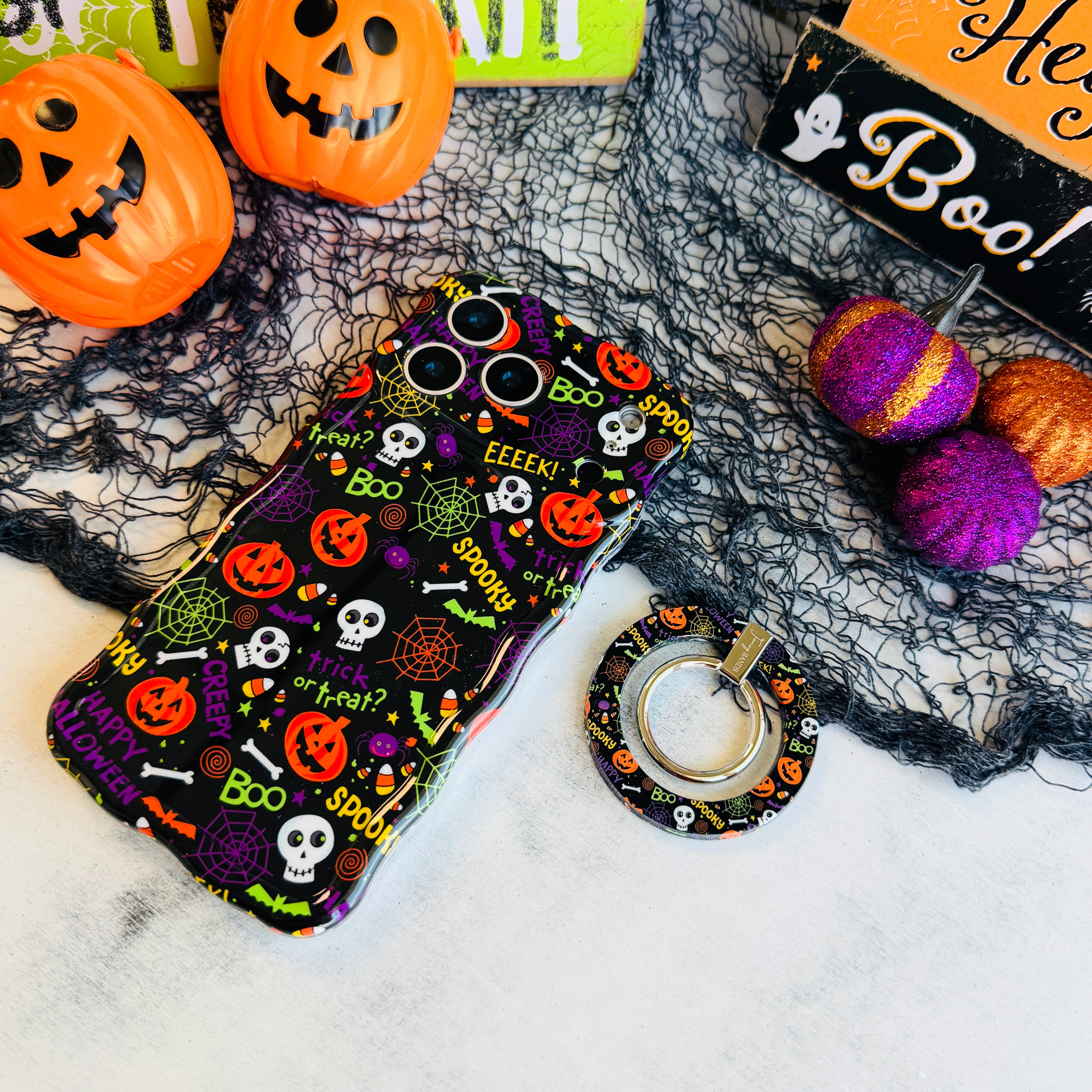 Spooky MagSafe grip ring lying beside the matching Wavy Luxe case, showing the black pumpkin and skull print on both.
