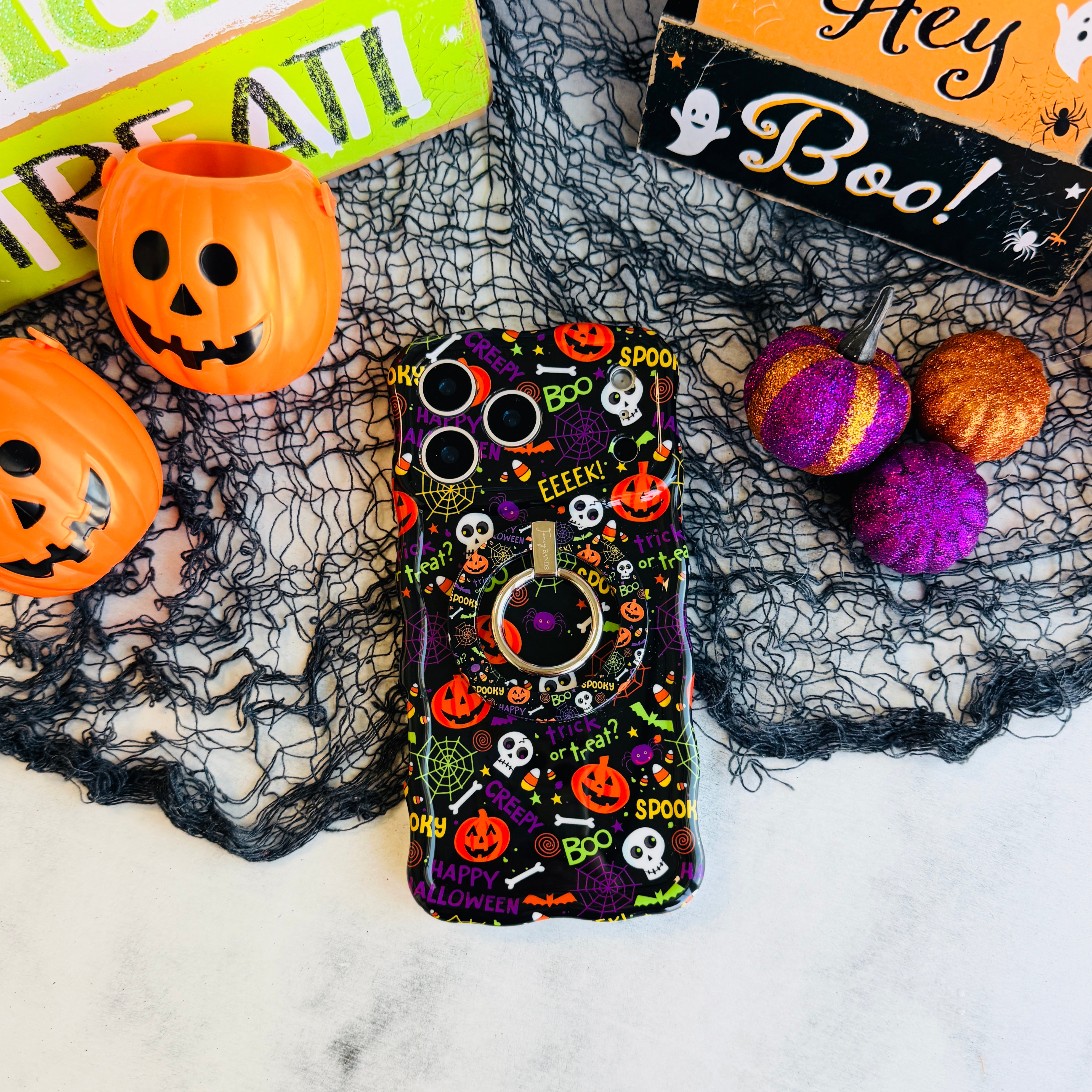 Spooky MagSafe grip ring attached to the matching Wavy Luxe case on black spiderweb mesh with an orange pail and a Hey Boo sign.