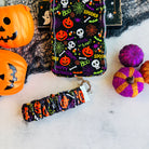 Spooky scrunchie phone wrist strap on marble beside the matching Wavy Luxe case, with an orange jack-o-lantern pail and felt pumpkins.