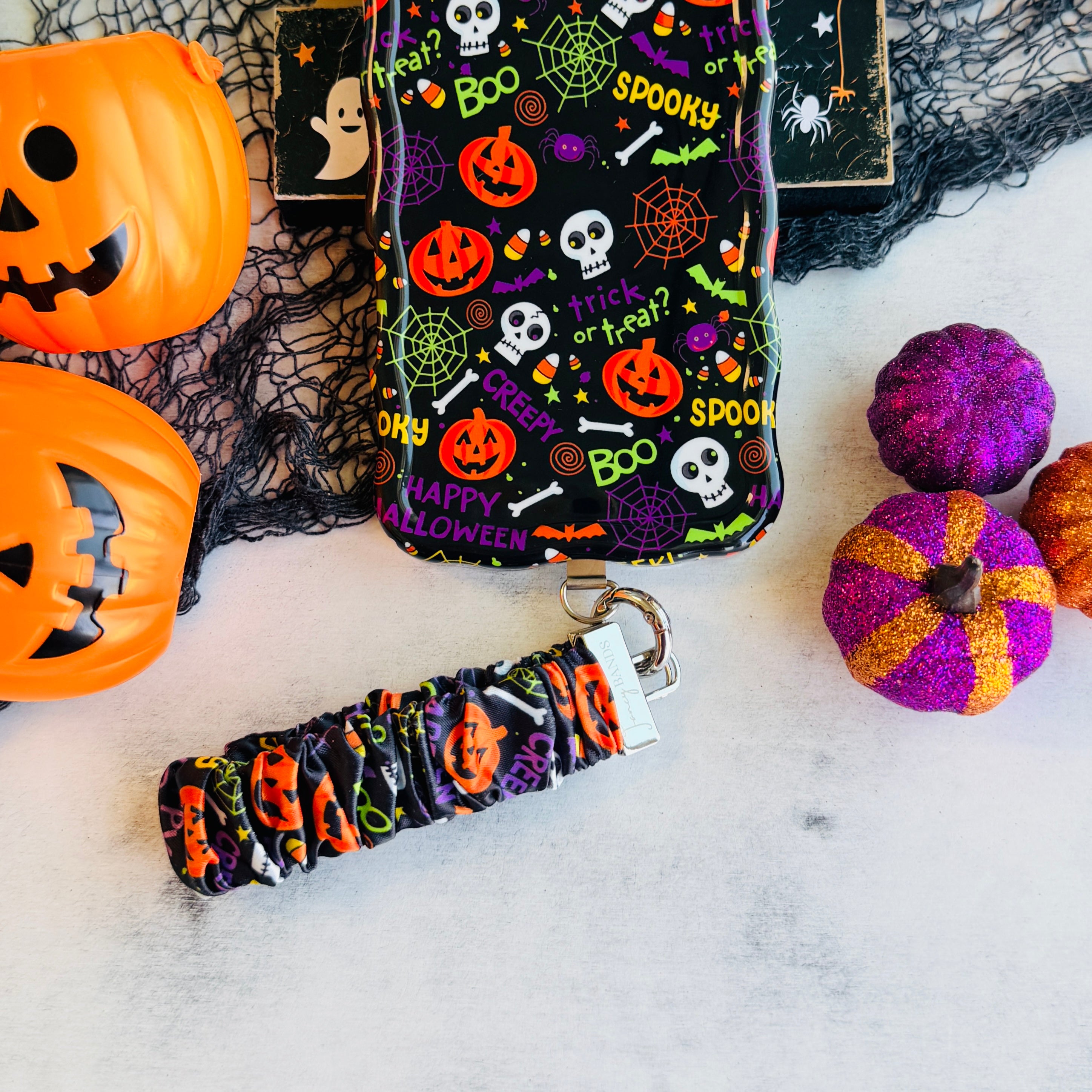 Spooky scrunchie phone wrist strap on marble beside the matching Wavy Luxe case, with an orange jack-o-lantern pail and felt pumpkins.
