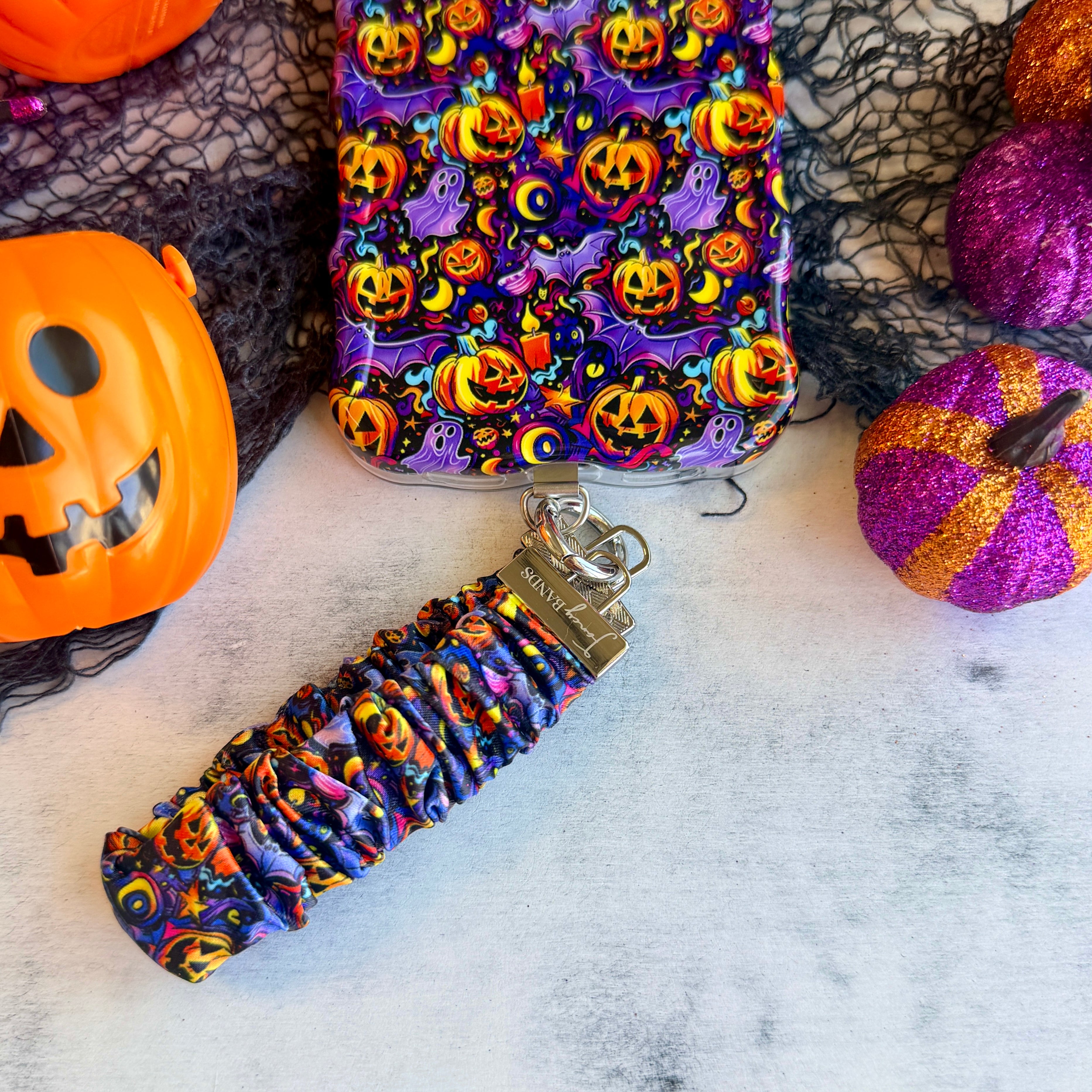 Jack-O-Lantern Jive scrunchie phone wrist strap on marble beside the matching Wavy Luxe case, with orange jack-o-lantern pails and glitter pumpkins.