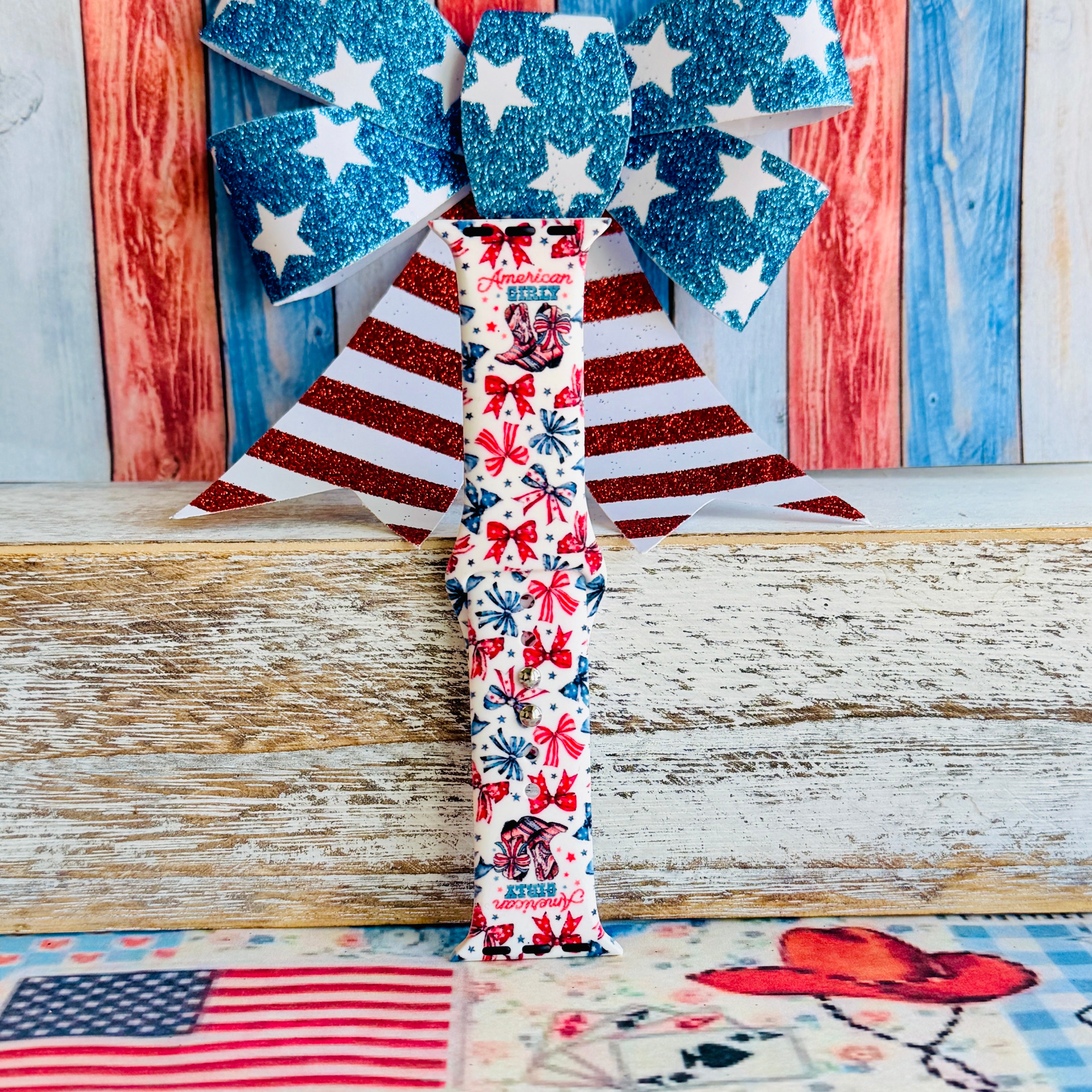 American Girly silicone Apple Watch band with red and blue coquette bows, mini cowboy boots, stars, and "American Girly" script on a white base, shown displayed and worn on Apple Watch against a patriotic 4th of July backdrop.