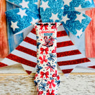 American Girly silicone Apple Watch band with red and blue coquette bows, mini cowboy boots, stars, and "American Girly" script on a white base, shown displayed and worn on Apple Watch against a patriotic 4th of July backdrop.