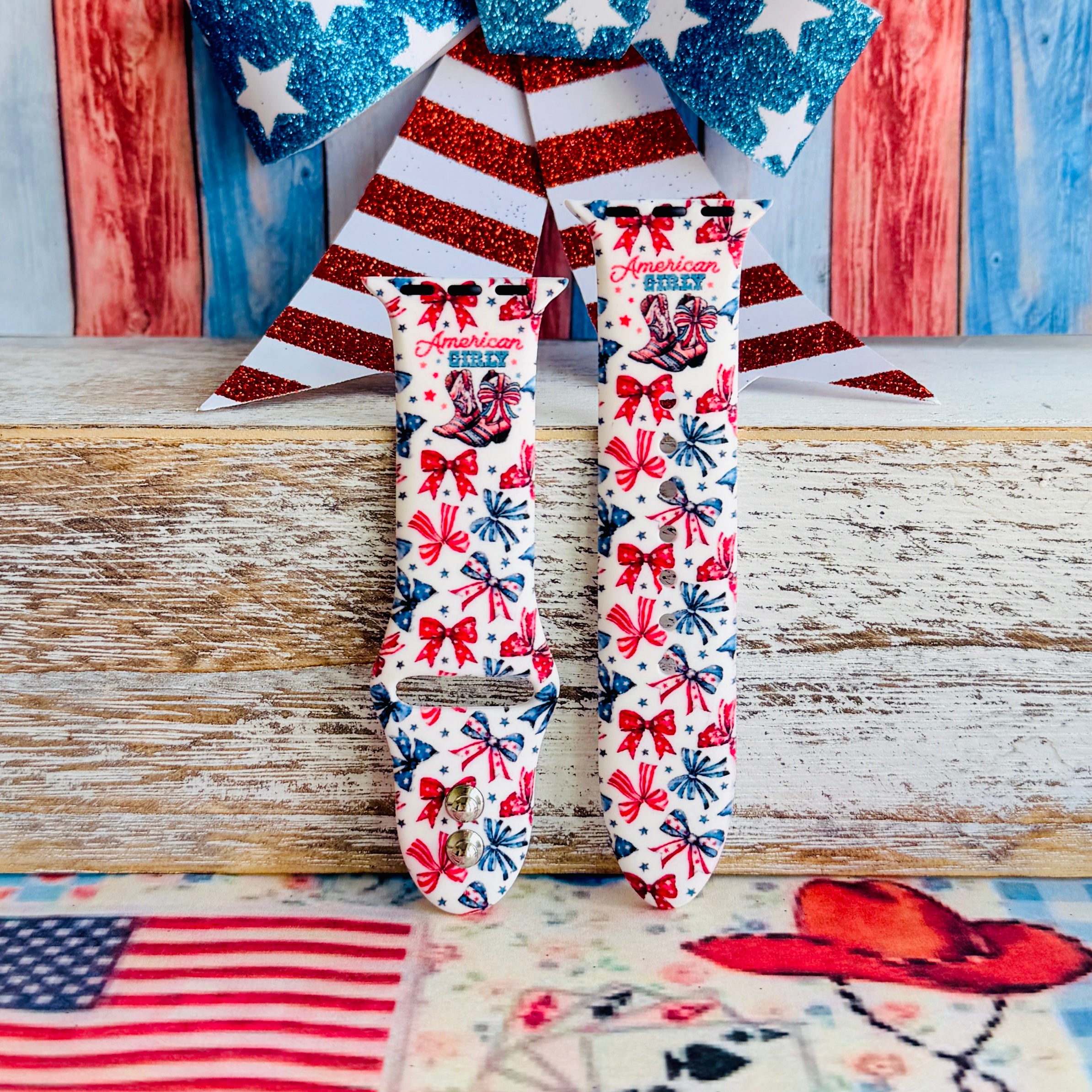 American Girly silicone Apple Watch band with red and blue coquette bows, mini cowboy boots, stars, and "American Girly" script on a white base, shown displayed and worn on Apple Watch against a patriotic 4th of July backdrop.
