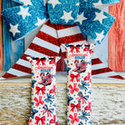 American Girly silicone Apple Watch band with red and blue coquette bows, mini cowboy boots, stars, and "American Girly" script on a white base, shown displayed and worn on Apple Watch against a patriotic 4th of July backdrop.
