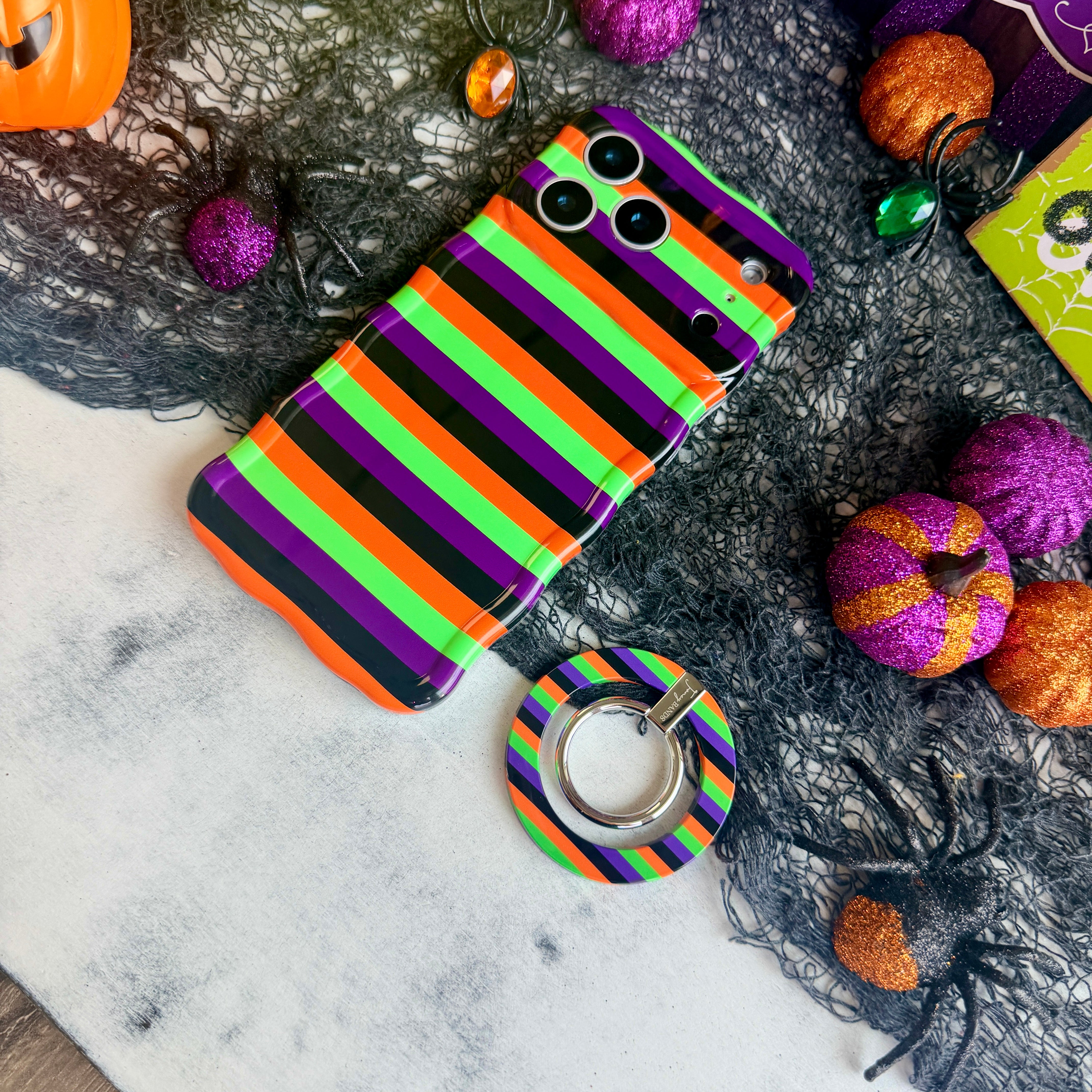 Main Witch Energy MagSafe grip ring lying beside the matching striped Wavy Luxe case, showing the black, purple, green and orange stripes on both.