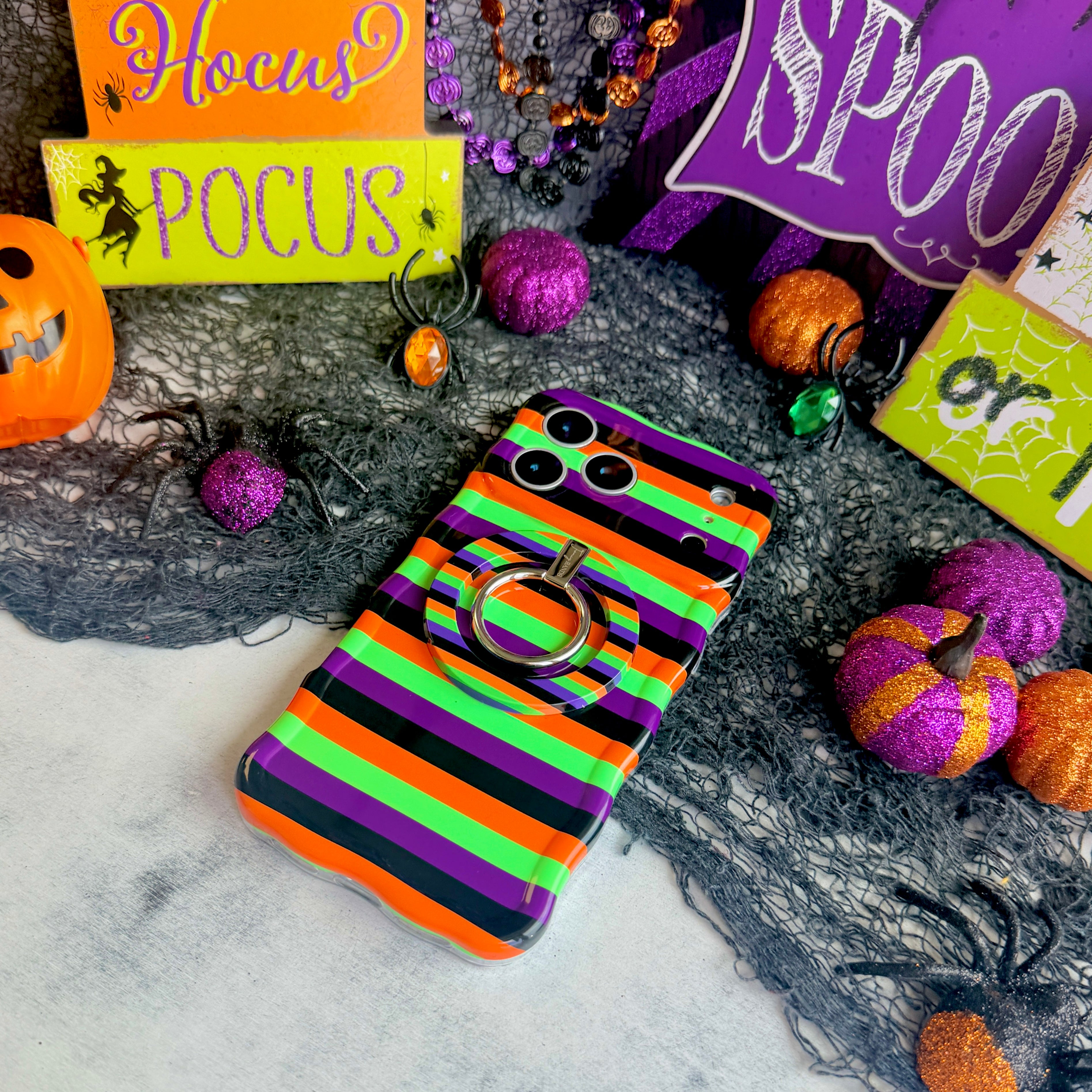 Main Witch Energy Wavy Luxe MagSafe iPhone case with the matching MagSafe grip ring attached, on a Halloween set with glitter pumpkins.