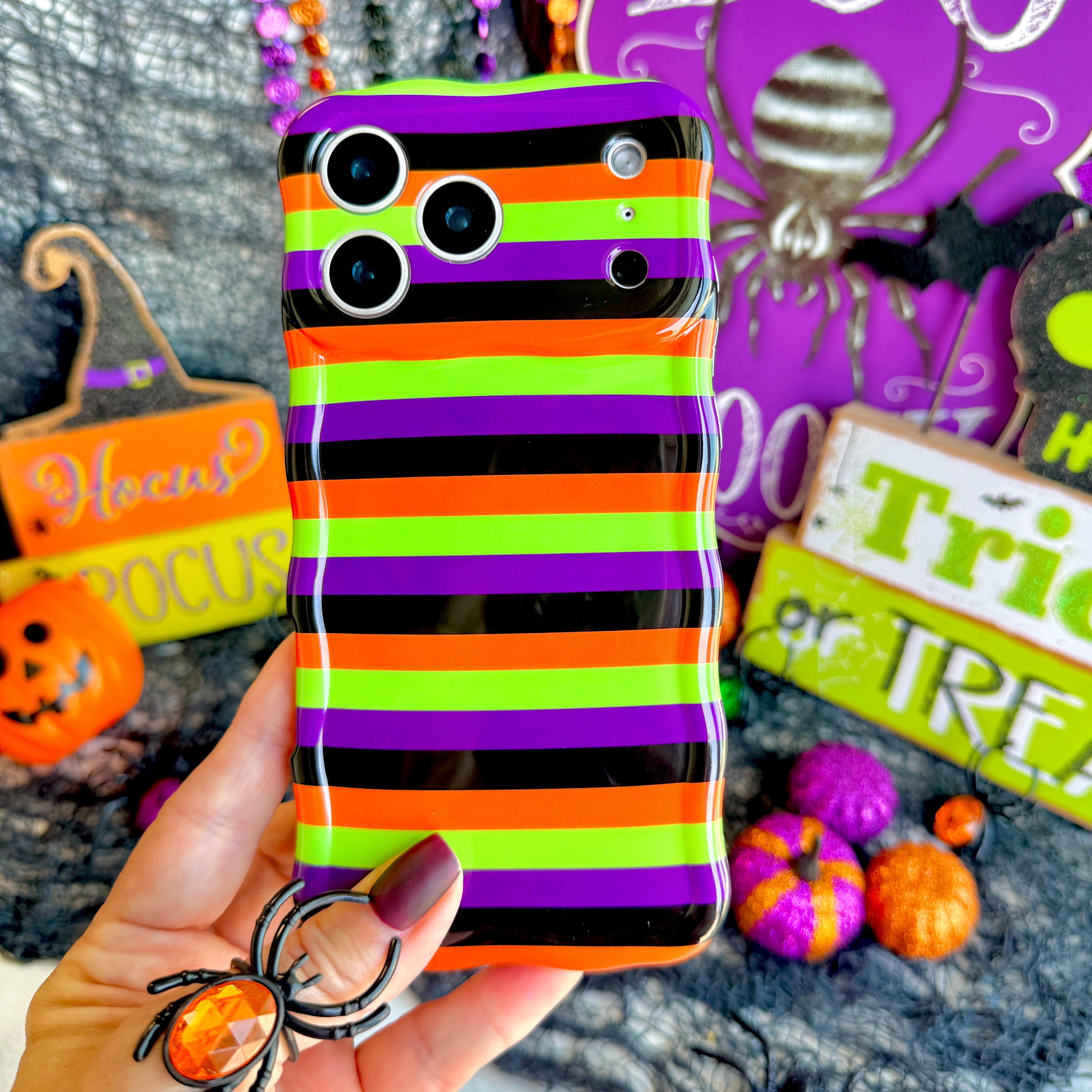 Main Witch Energy Wavy Luxe MagSafe iPhone case held in hand, striped in black, purple, neon green and orange, with a Hocus Pocus sign behind.