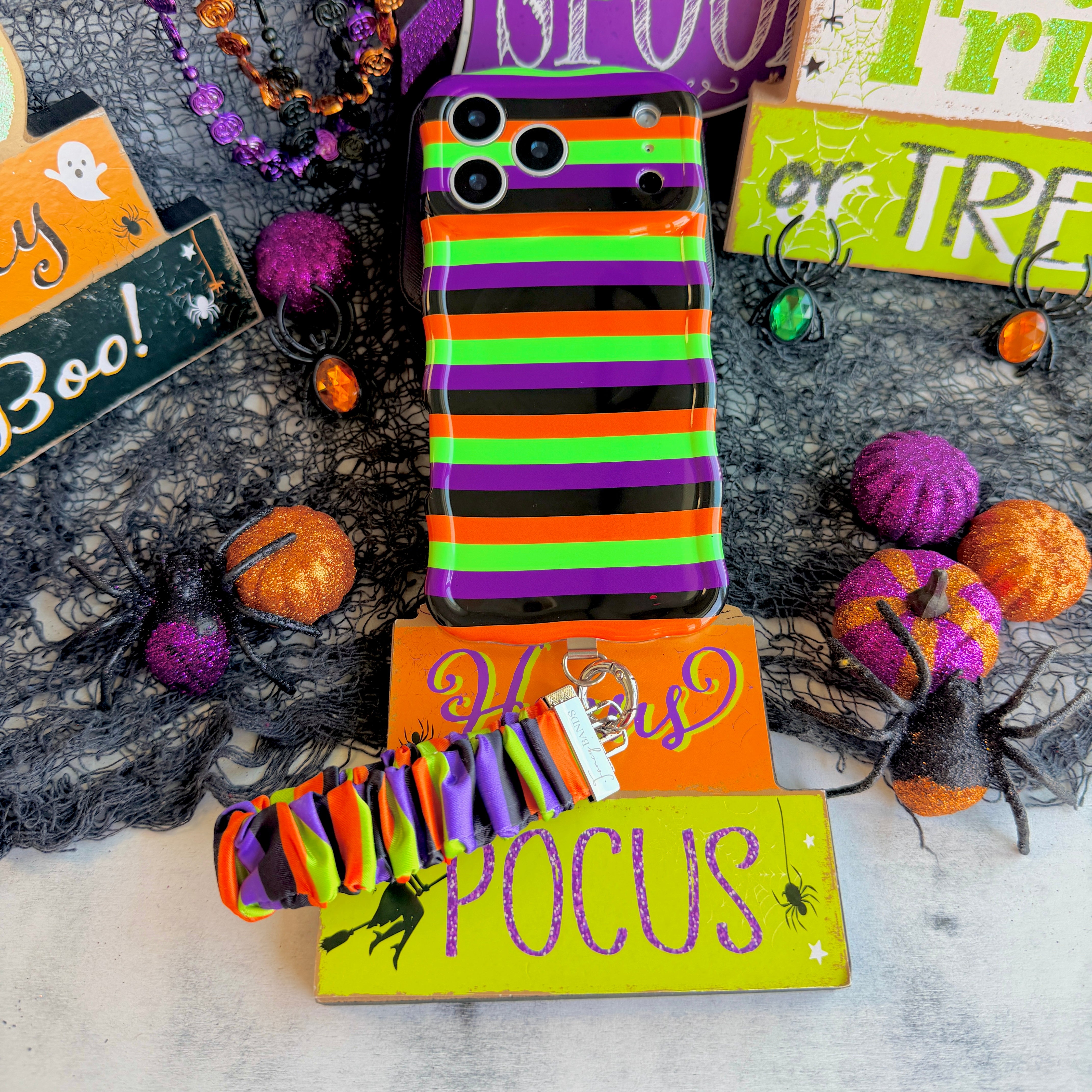Main Witch Energy scrunchie phone wrist strap and matching striped Wavy Luxe case shown together on a Hocus Pocus sign.