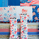 Party In The USA silicone Apple Watch band with fireworks, balloons, cupcakes, flag bunting, and stars in red, white, and blue on a cream base, shown displayed and worn on Apple Watch against a patriotic 4th of July backdrop.