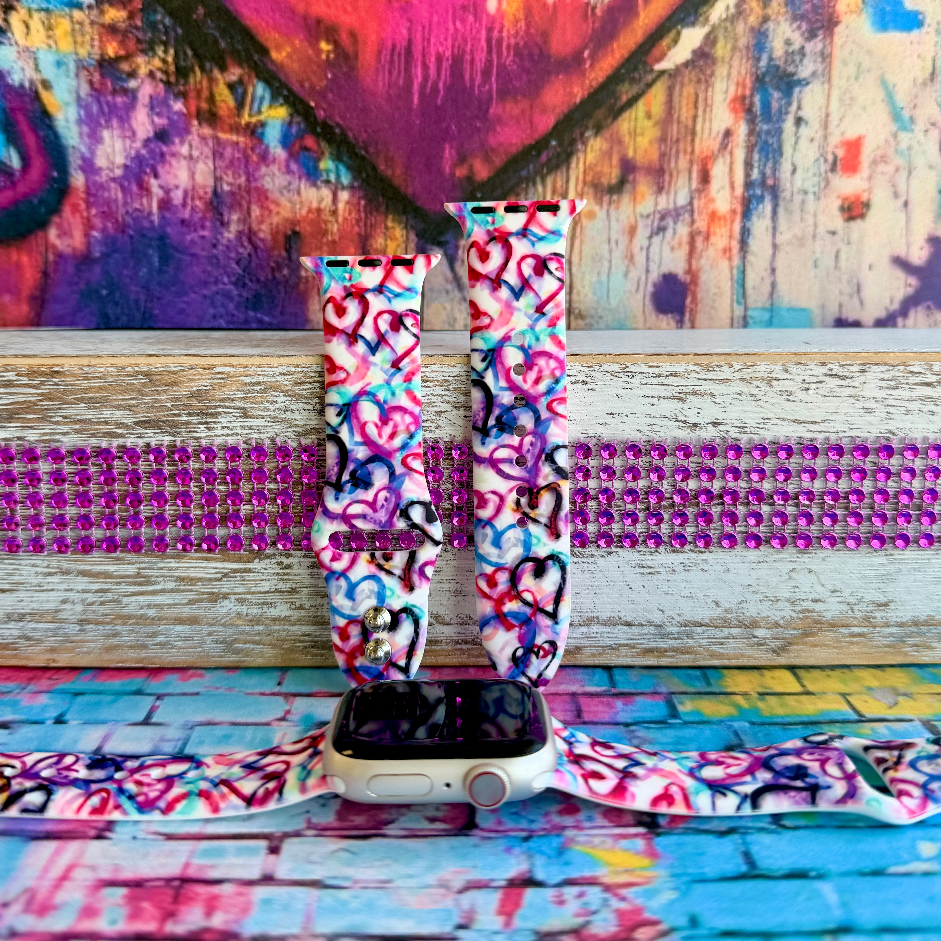 Love Out Loud silicone Apple Watch band with hand-drawn graffiti-style hearts in pink, magenta, blue, teal, red, and black on a white base, shown displayed and worn on Apple Watch against a colorful street art backdrop.